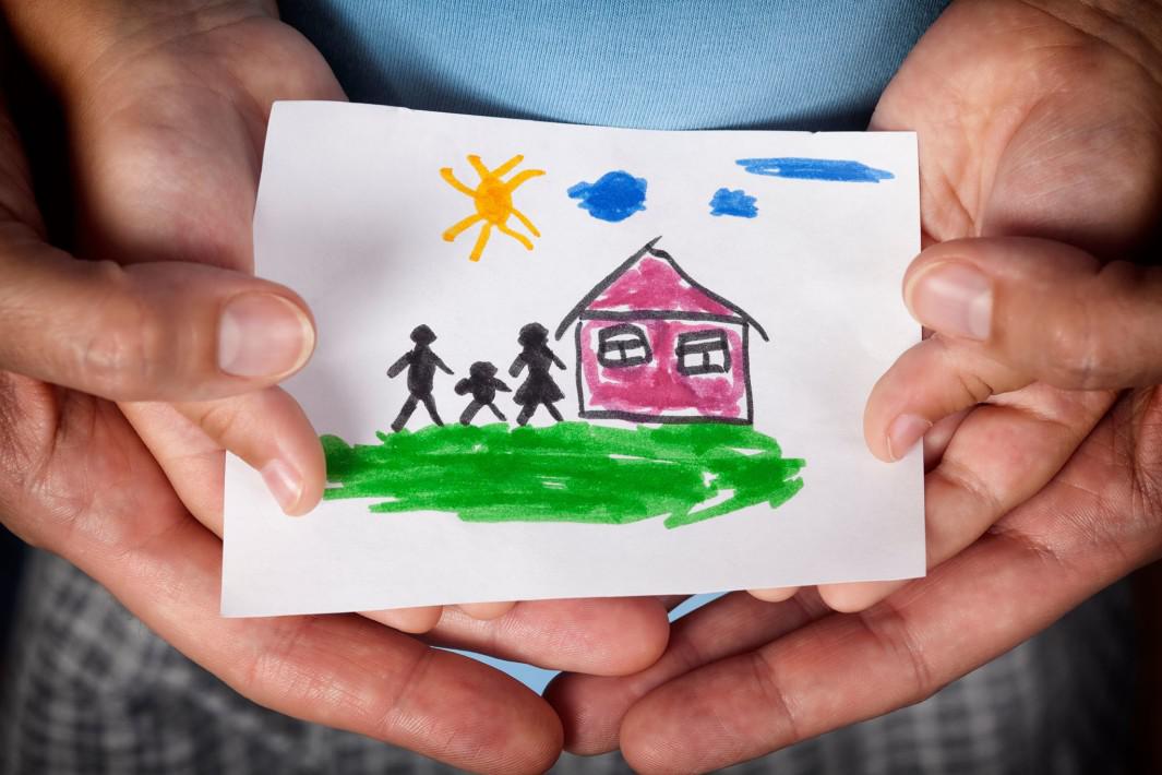#ParaCegoVer: as mãos de uma criança e de um adulto, juntas, seguram o desenho de uma família em frente à uma casa rosa em um dia de sol.