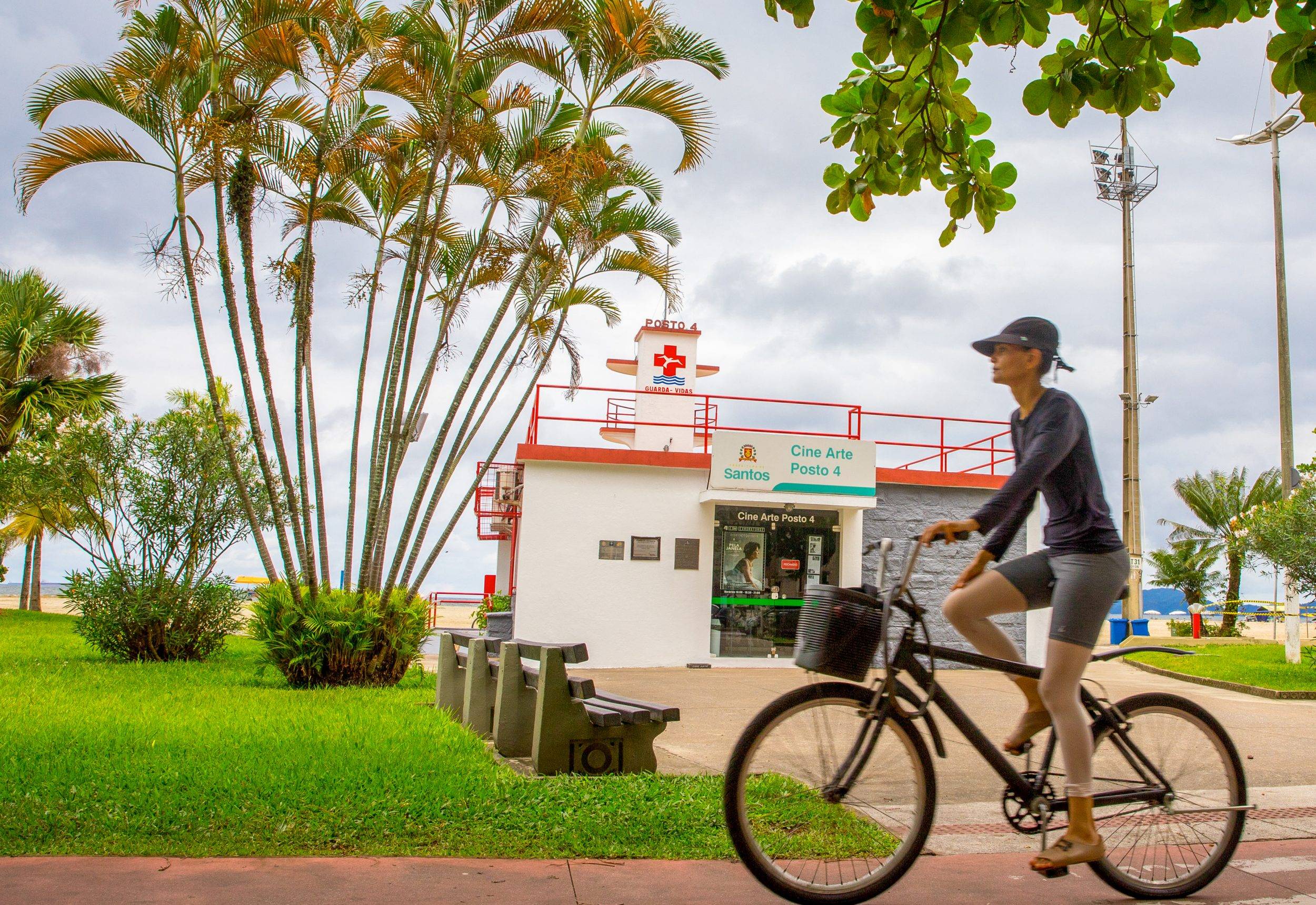 Mulher de boné e roupa escura pedalando em ciclovia, com coqueiros e posto de salvamento ao fundo em Santos.