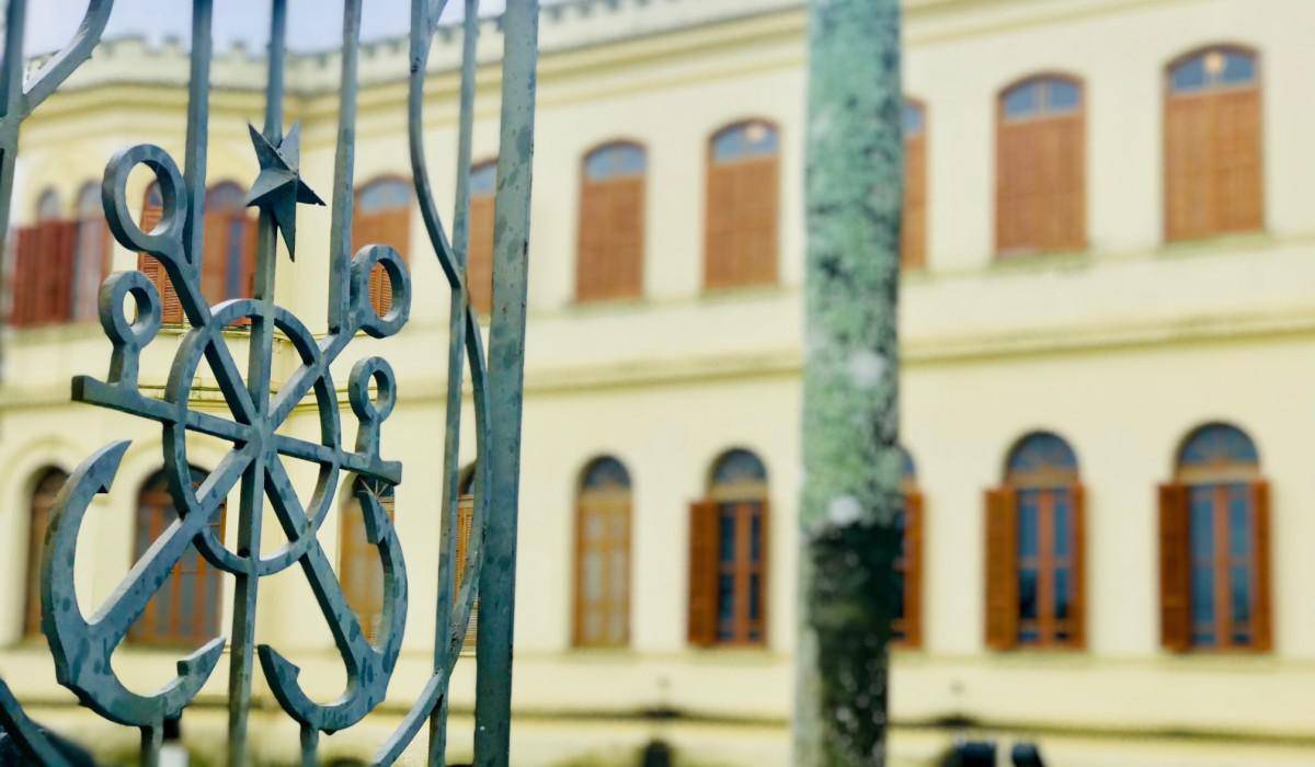 Grade de ferro com âncora e leme em primeiro plano, em frente a um edifício histórico amarelo com janelas arqueadas.