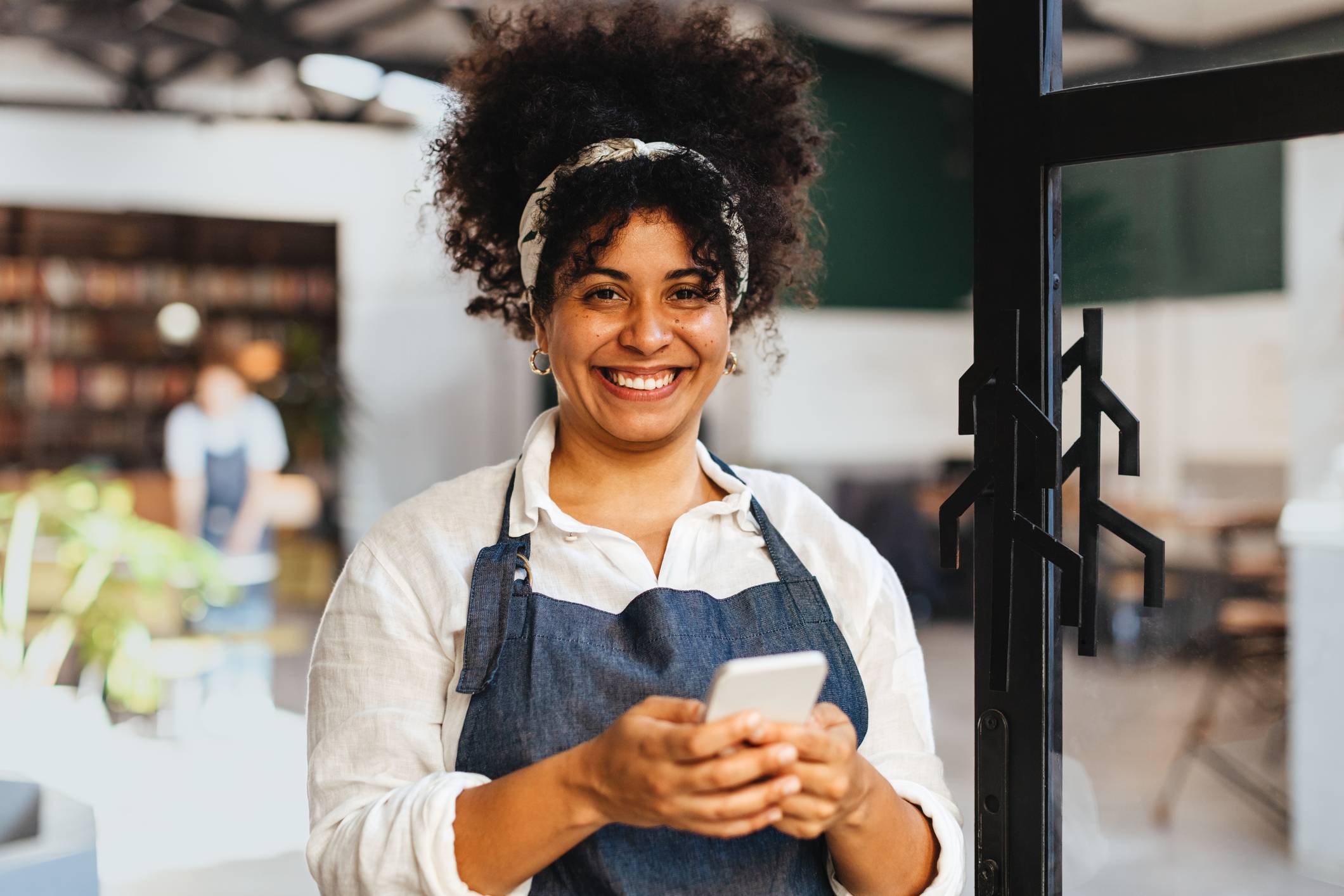 Mulher negra sorridente de cabelo cacheado, avental, segurando celular na entrada de café.