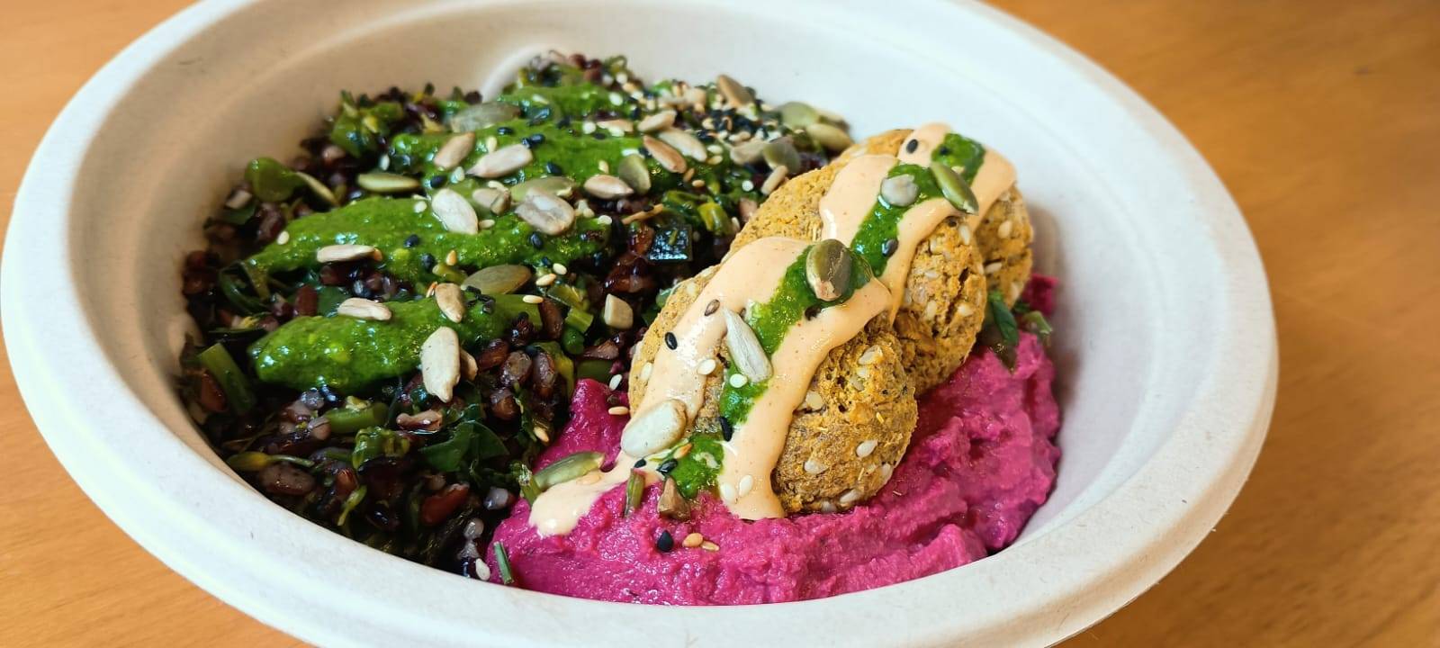 Tigela de refeição colorida com arroz preto, vegetais, pesto, homus de beterraba e falafel, coberta com molhos e sementes.
