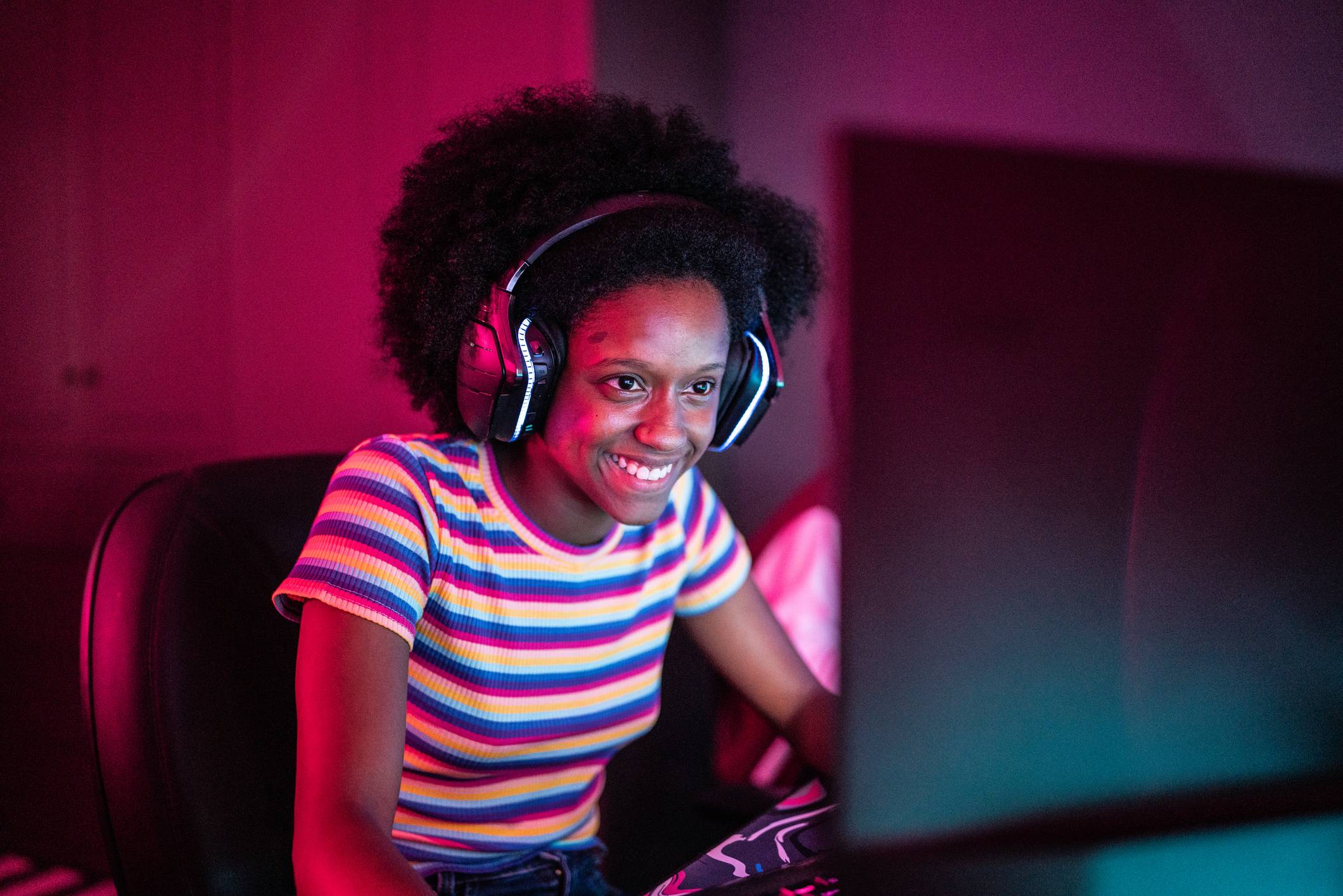 Jovem mulher negra com cabelo crespo e fones de ouvido sorri enquanto usa computador em ambiente com luzes neon.