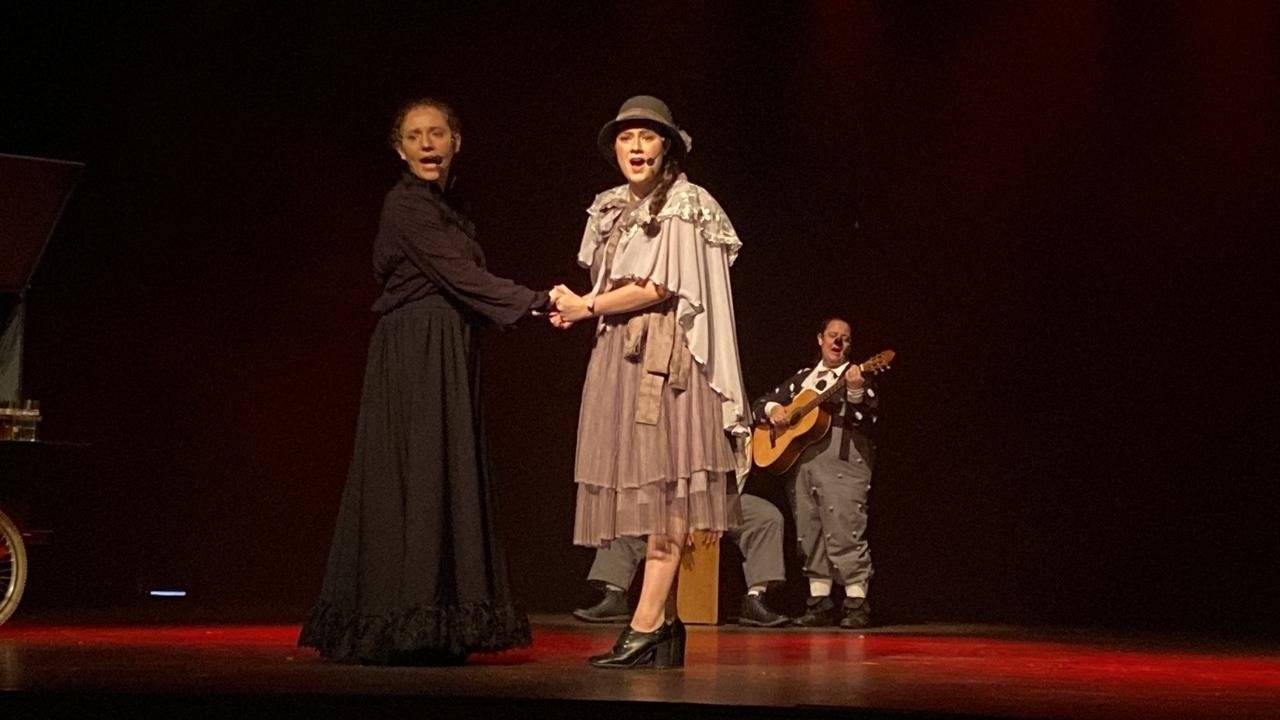 Duas mulheres cantando de mãos dadas no palco, uma de vestido escuro e outra de chapéu, com músico de guitarra ao fundo.