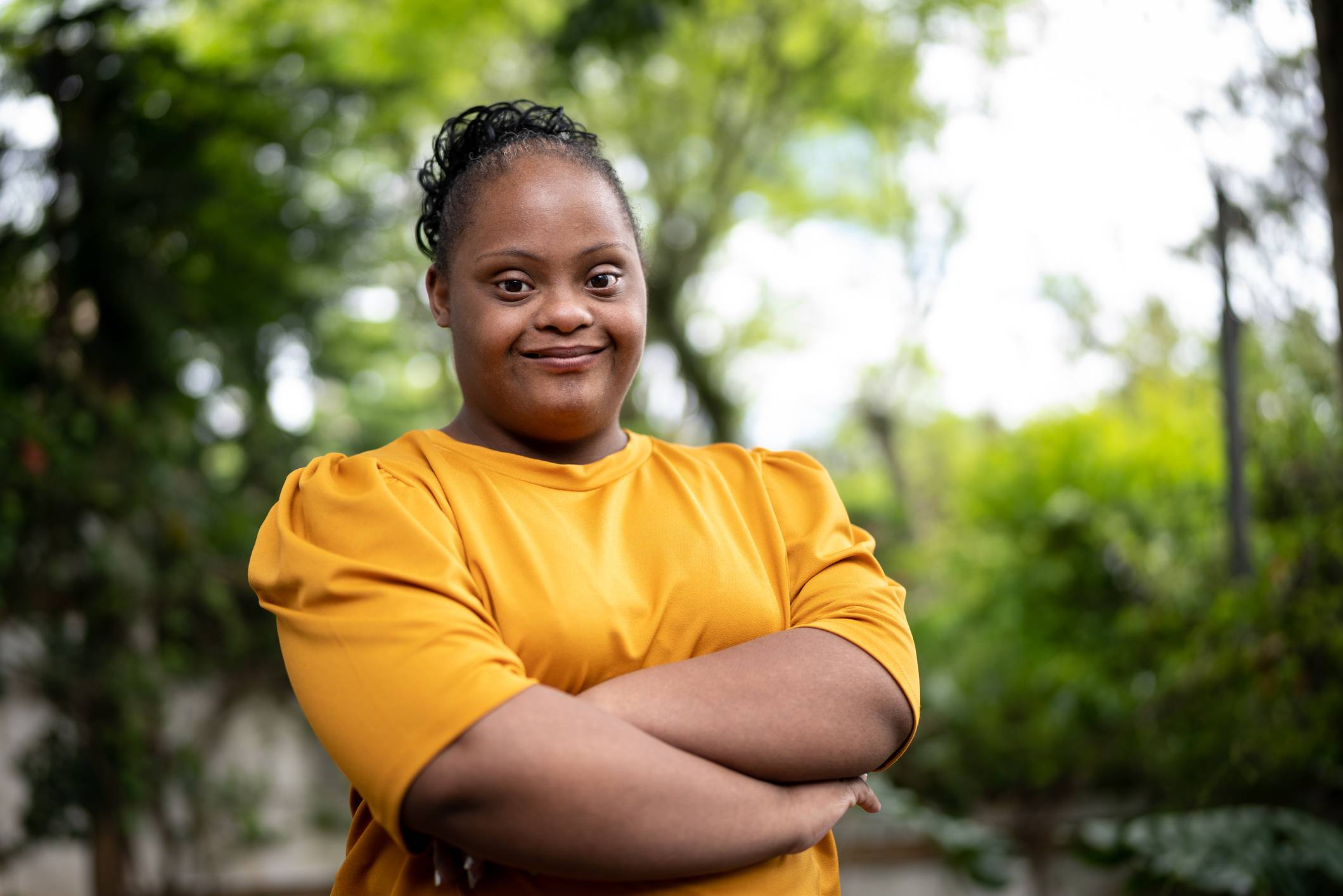 Mulher negra com síndrome de Down sorrindo, braços cruzados, blusa amarela, em ambiente externo com fundo verde borrado.
