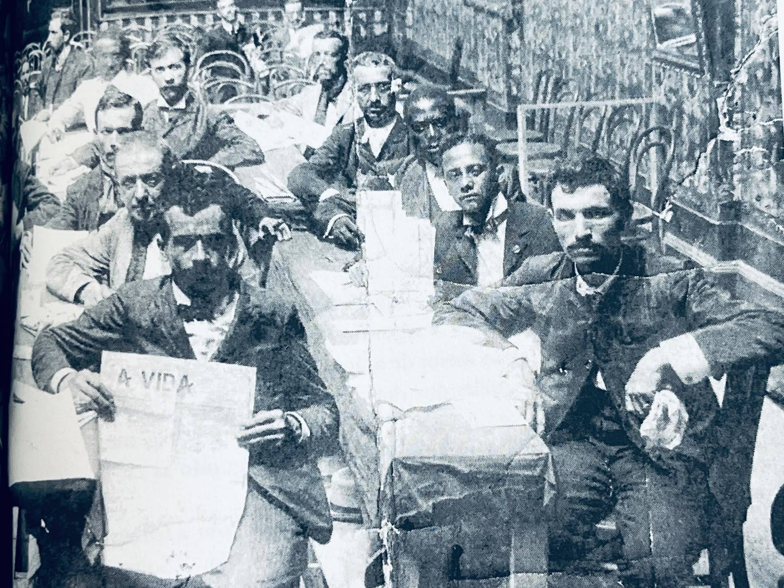 Grupo diverso de homens em trajes formais sentados em mesas de um café antigo, um deles lendo o jornal 'A VIDA'.