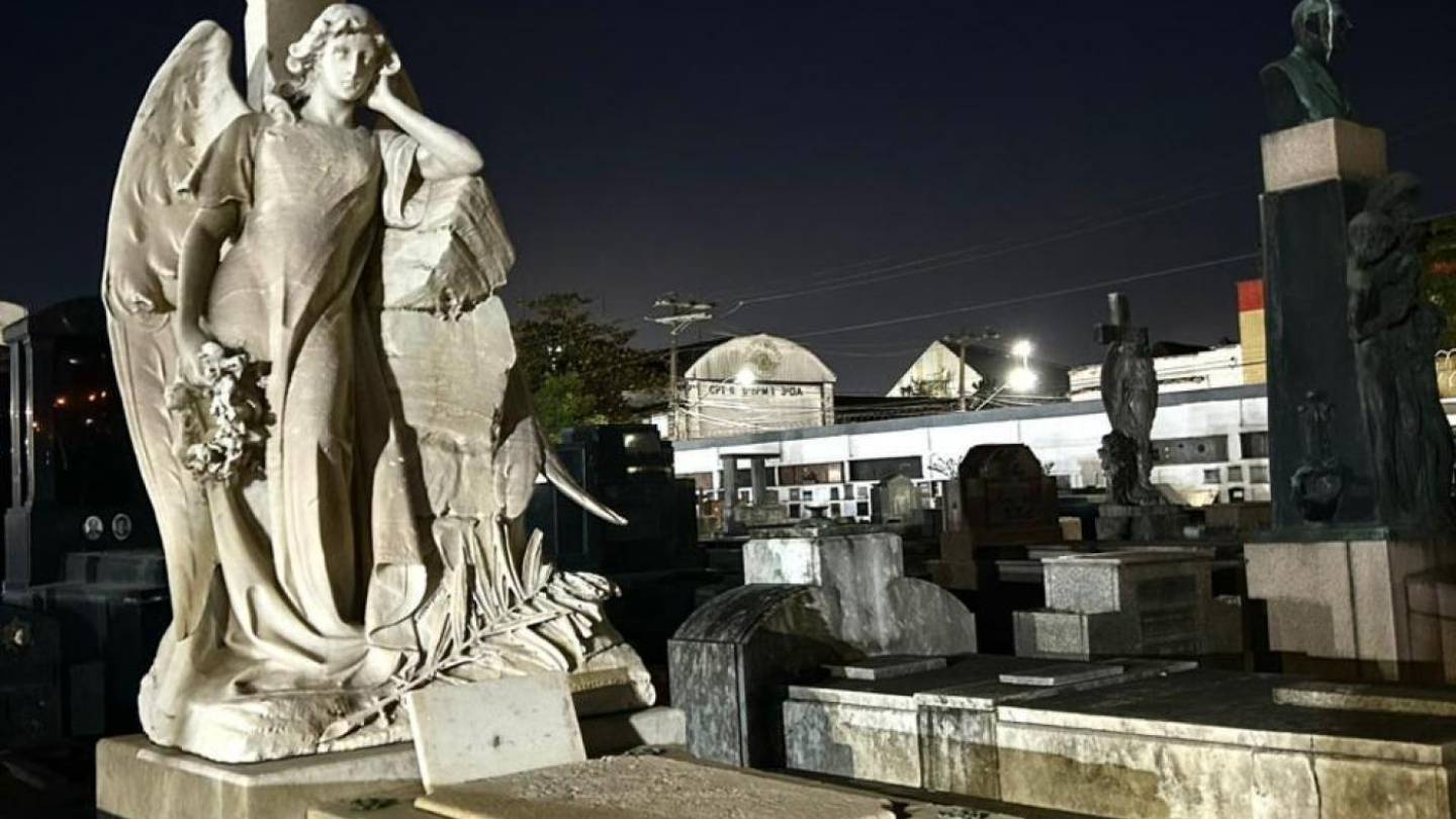 Estátua de anjo com asas segurando flores em cemitério à noite, com lápides e prédios iluminados ao fundo.