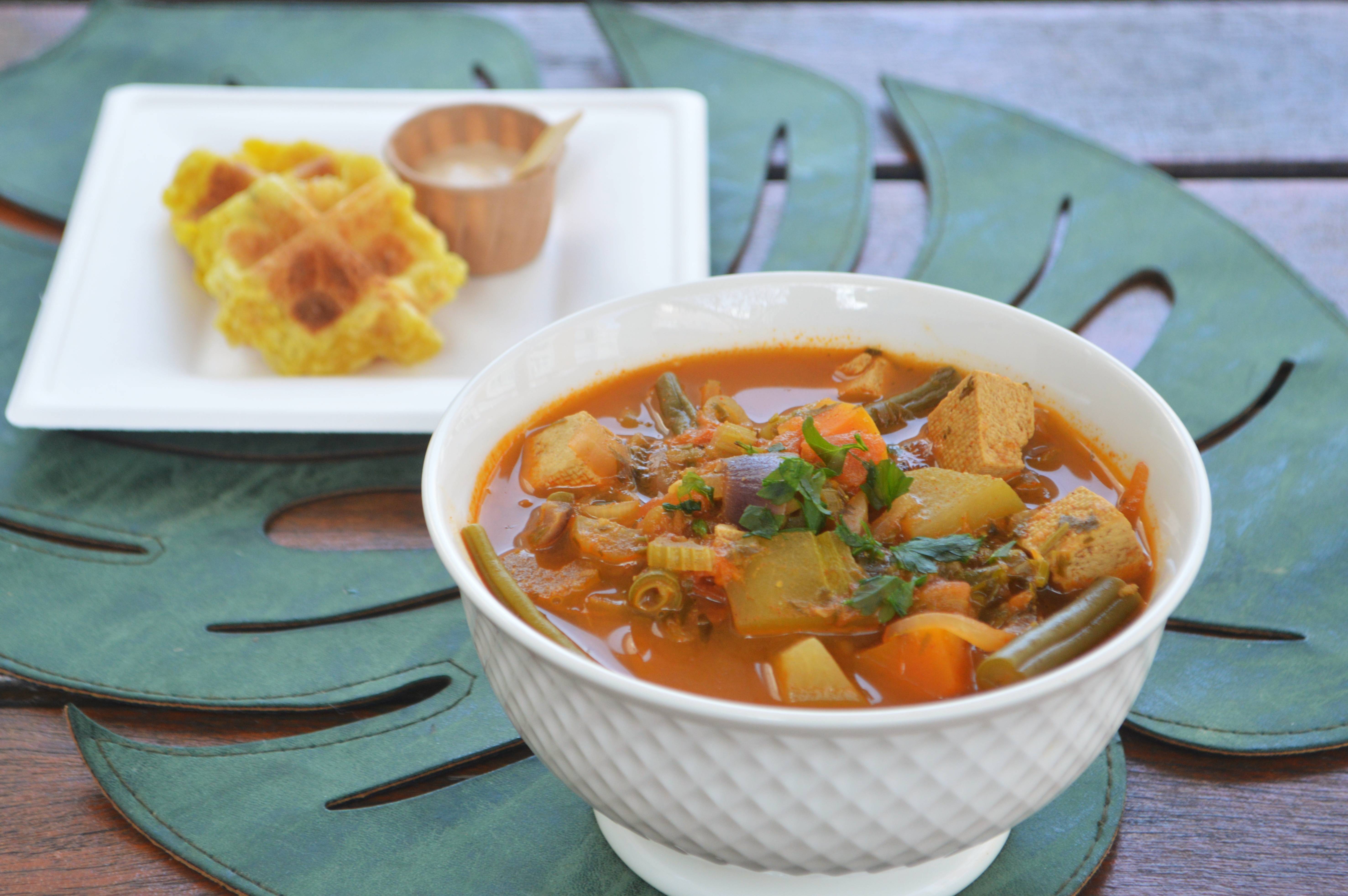 Tigela de ensopado de vegetais com tofu e caldo avermelhado, acompanhada de waffles salgados e placemats em forma de folha