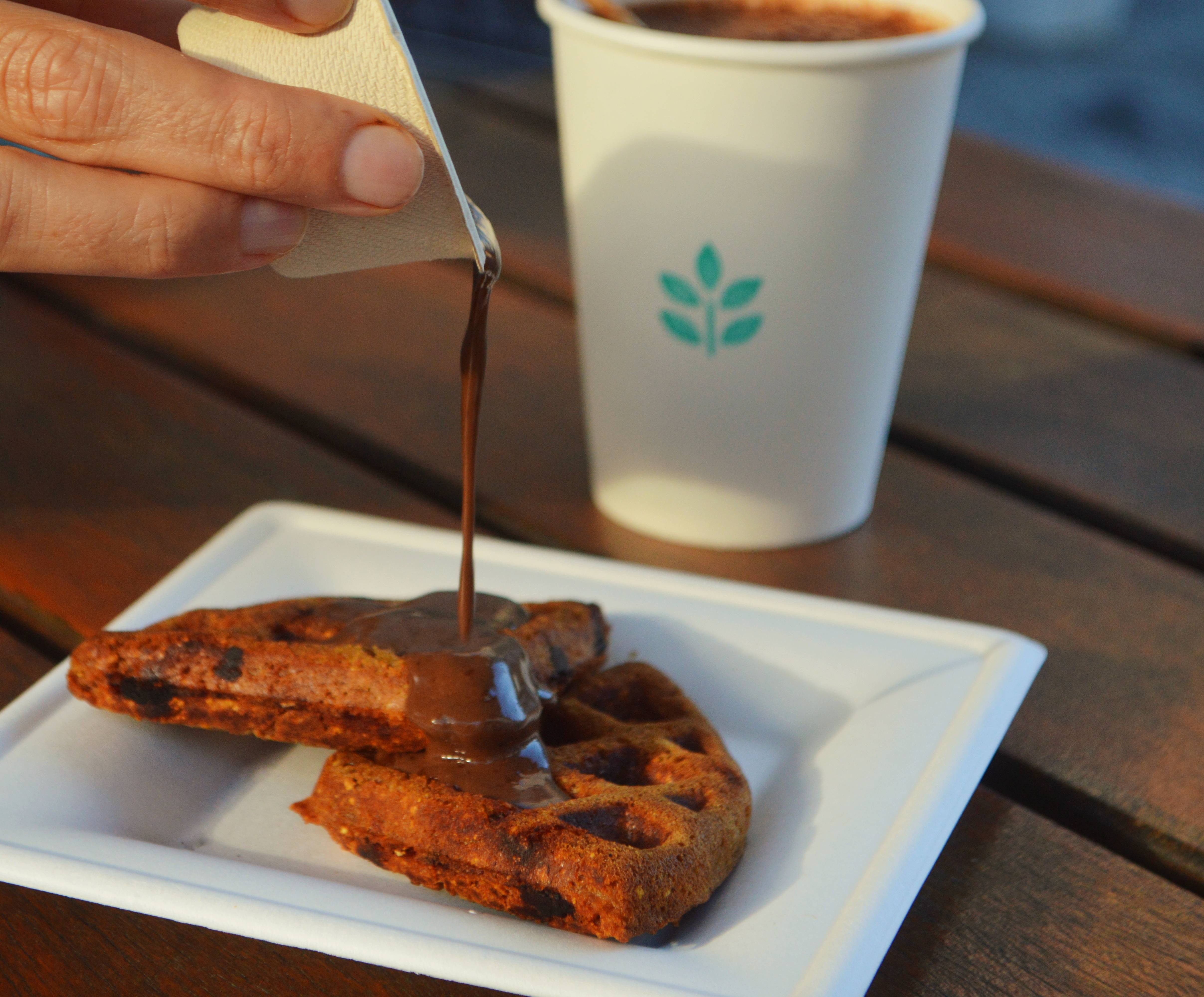 Mão derramando calda de chocolate escura sobre dois waffles crocantes em um prato branco, com copo de bebida quente ao fundo.