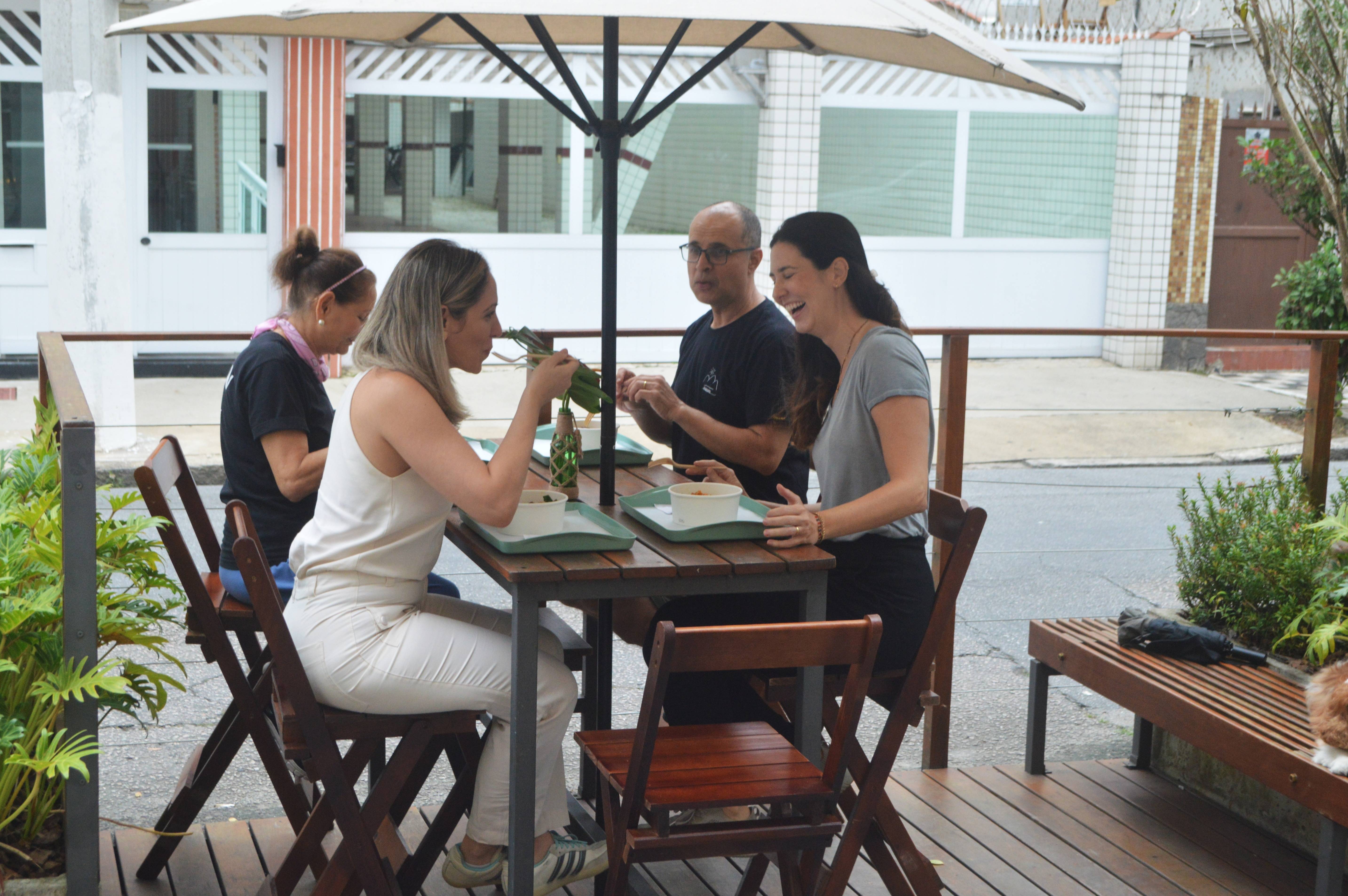 Pessoas comendo e rindo em mesa de madeira ao ar livre sob guarda-sol, desfrutando de uma refeição em restaurante casual.