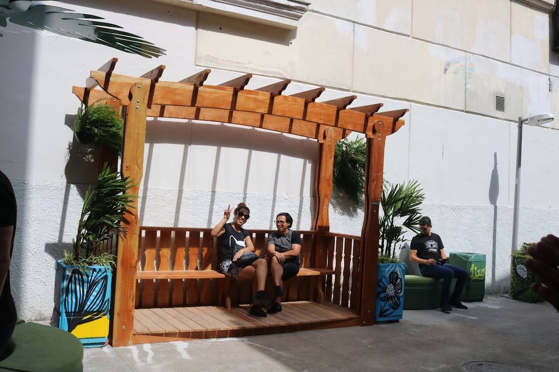 Pessoas relaxando em banco de madeira sob pérgola, com vasos de plantas decorados e um homem sentado em banco verde.