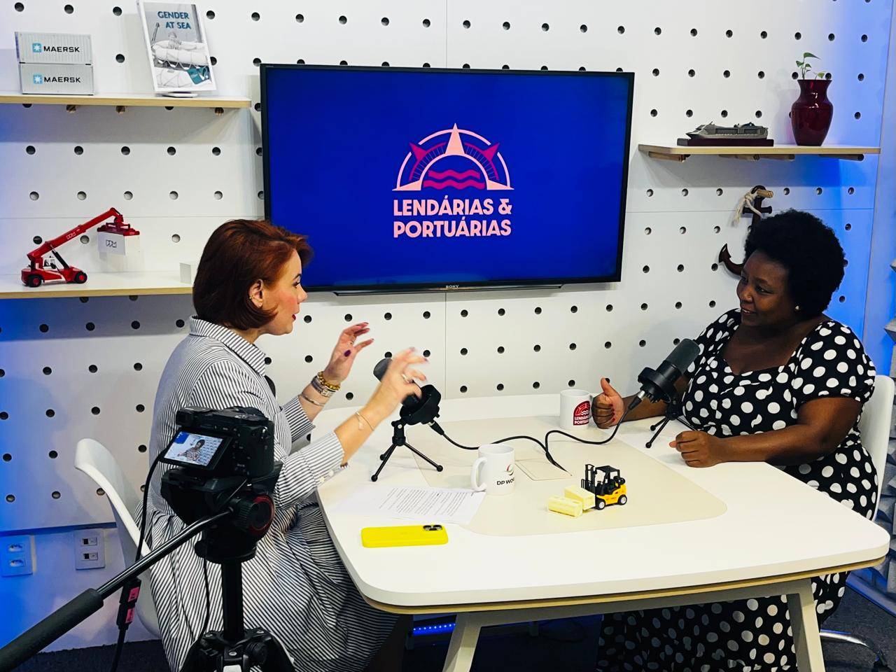 Duas mulheres conversando e gesticulando em um estúdio de podcast com uma tela ao fundo exibindo 'Lendárias & Portuárias', rodeadas por miniaturas de equipamentos portuários e marítimos.