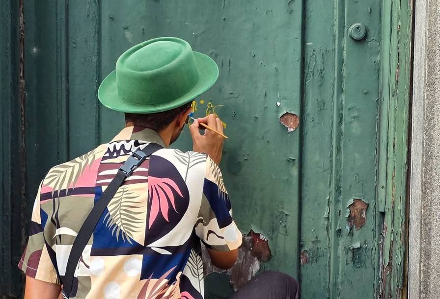Texto Alternativo (Alt): Homem de chapéu verde e camisa estampada pintando figuras amarelas em porta verde descascada.
Título da Imagem: Artista de chapéu verde pintando porta de madeira desgastada
Legenda: Um artista misterioso, usando um chapéu verde vibrante e uma camisa estampada, adiciona toques de cor e arte a uma porta de madeira desgastada.
Descrição: Imagem capturada por trás de um artista não identificado, com um chapéu verde marcante e uma camisa de padrão tropical ou abstrato, enquanto ele cuidadosamente pinta figuras ou símbolos em amarelo em uma velha porta de madeira verde. A porta exibe sinais visíveis de desgaste, com a tinta descascada revelando a textura da madeira por baixo. A cena evoca um senso de criatividade urbana e o contraste entre o novo e o antigo.