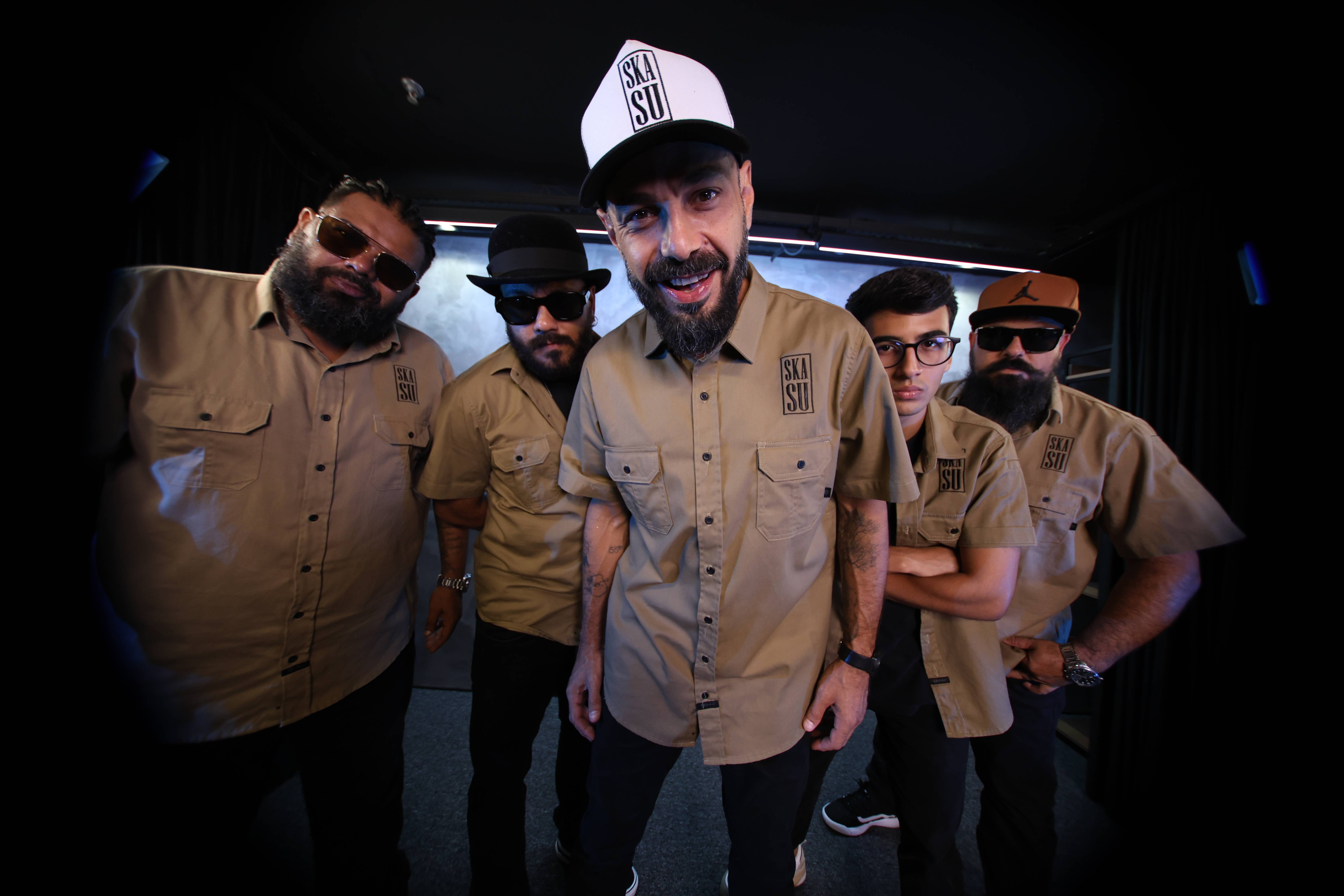 Cinco membros da banda Skasu, com camisas cáqui combinando e chapéus, posam sorrindo em um ambiente escuro.