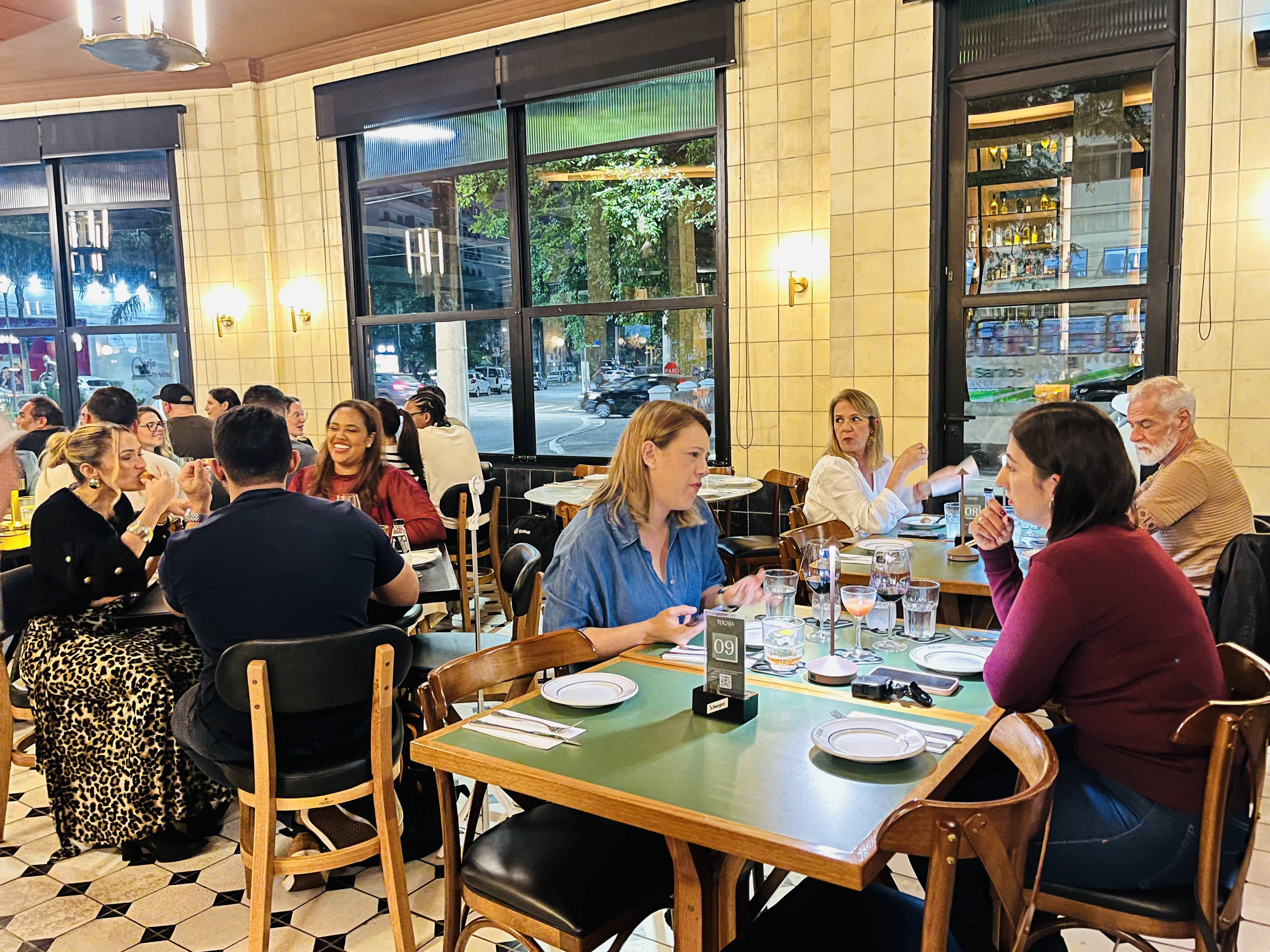 Pessoas jantando em um restaurante com grandes janelas, mesas de madeira e azulejos claros, ambiente noturno.