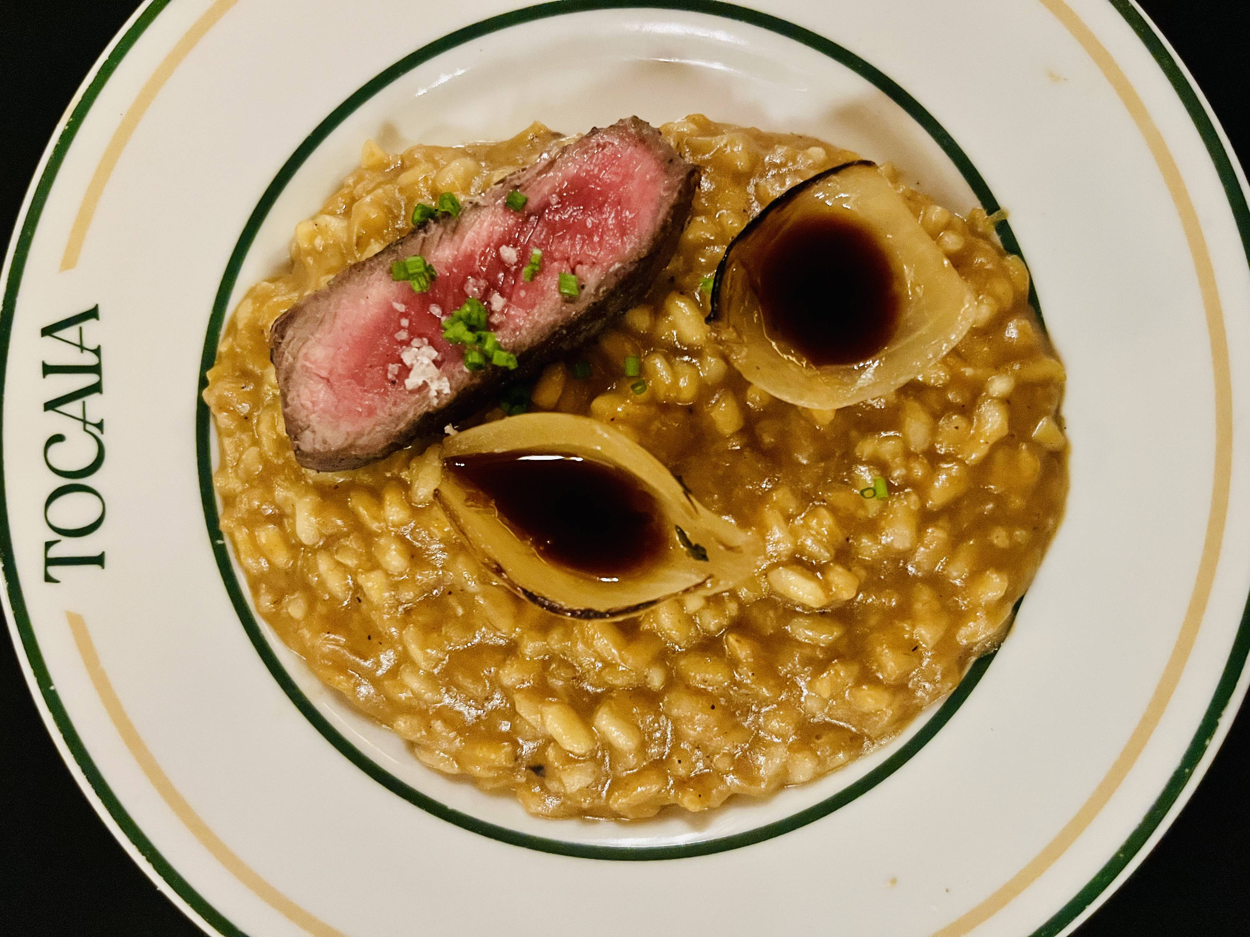 Risoto cremoso com bife malpassado, sal grosso, cebolinha e cebolas caramelizadas com molho, servido em prato Tocaia.