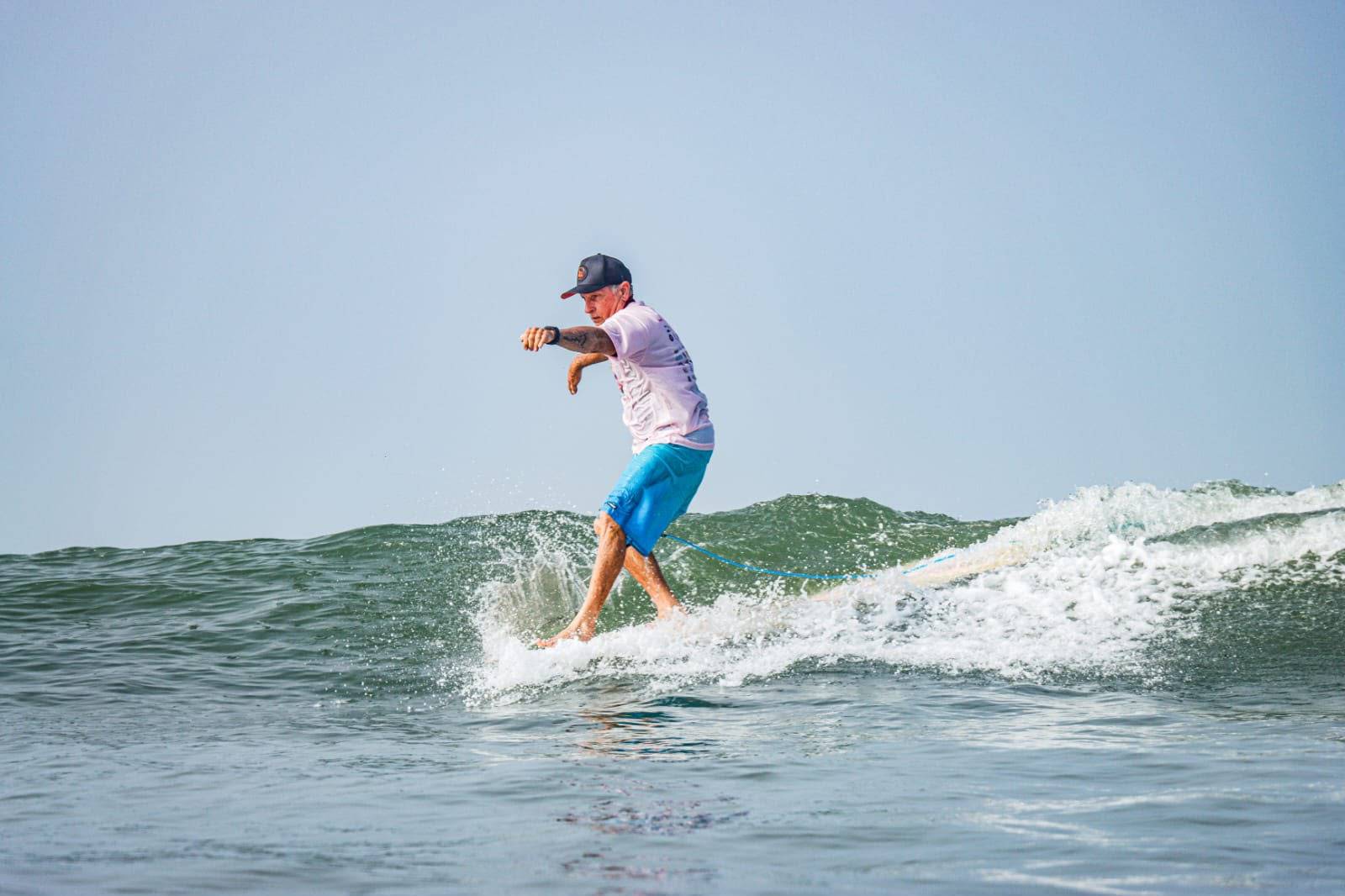 Homem de boné, camiseta clara e bermuda azul surfando em uma onda com espuma. Ele está equilibrando-se na prancha.