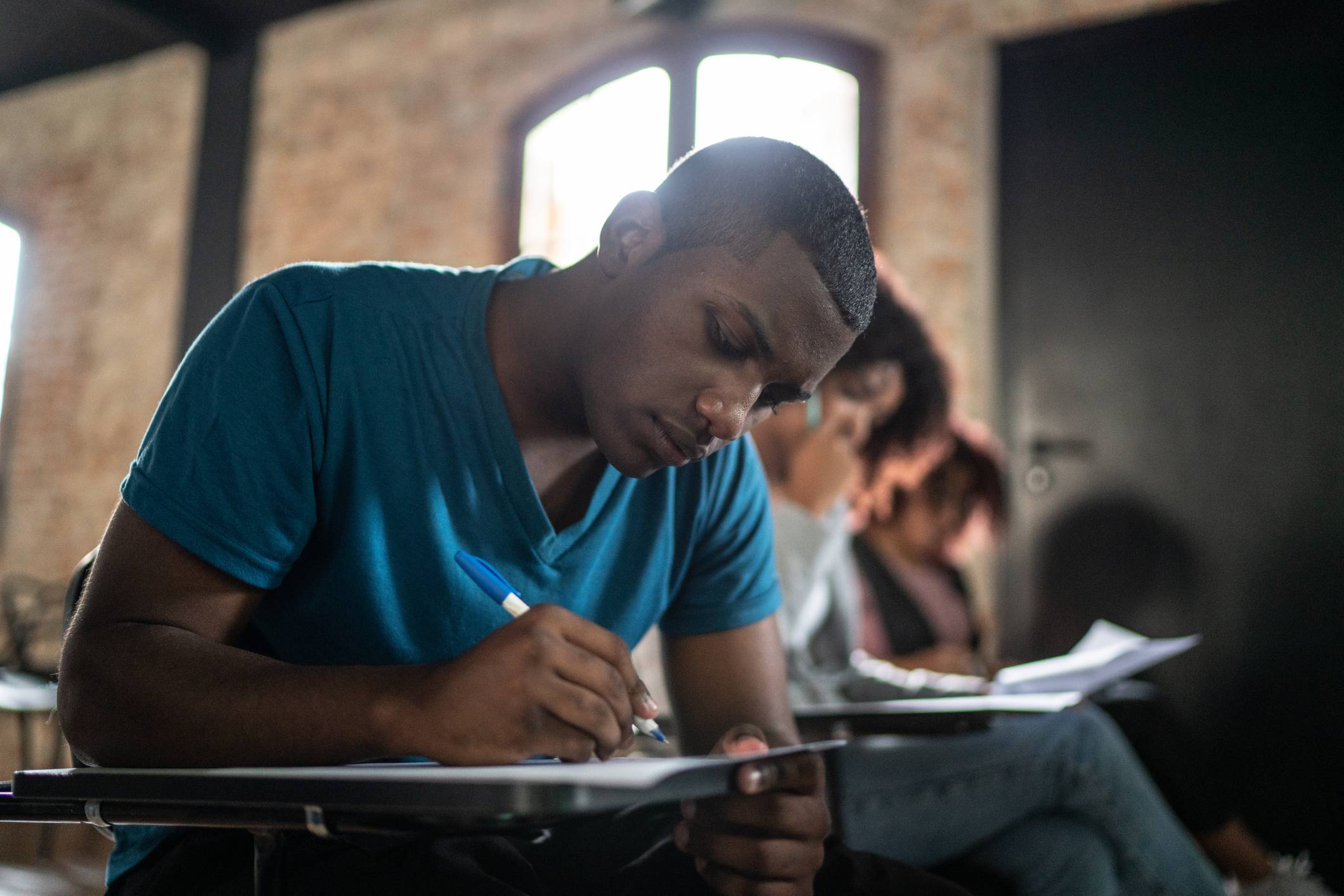 Jovem estudante negro escrevendo concentrado com caneta azul em papel sobre uma carteira escolar.