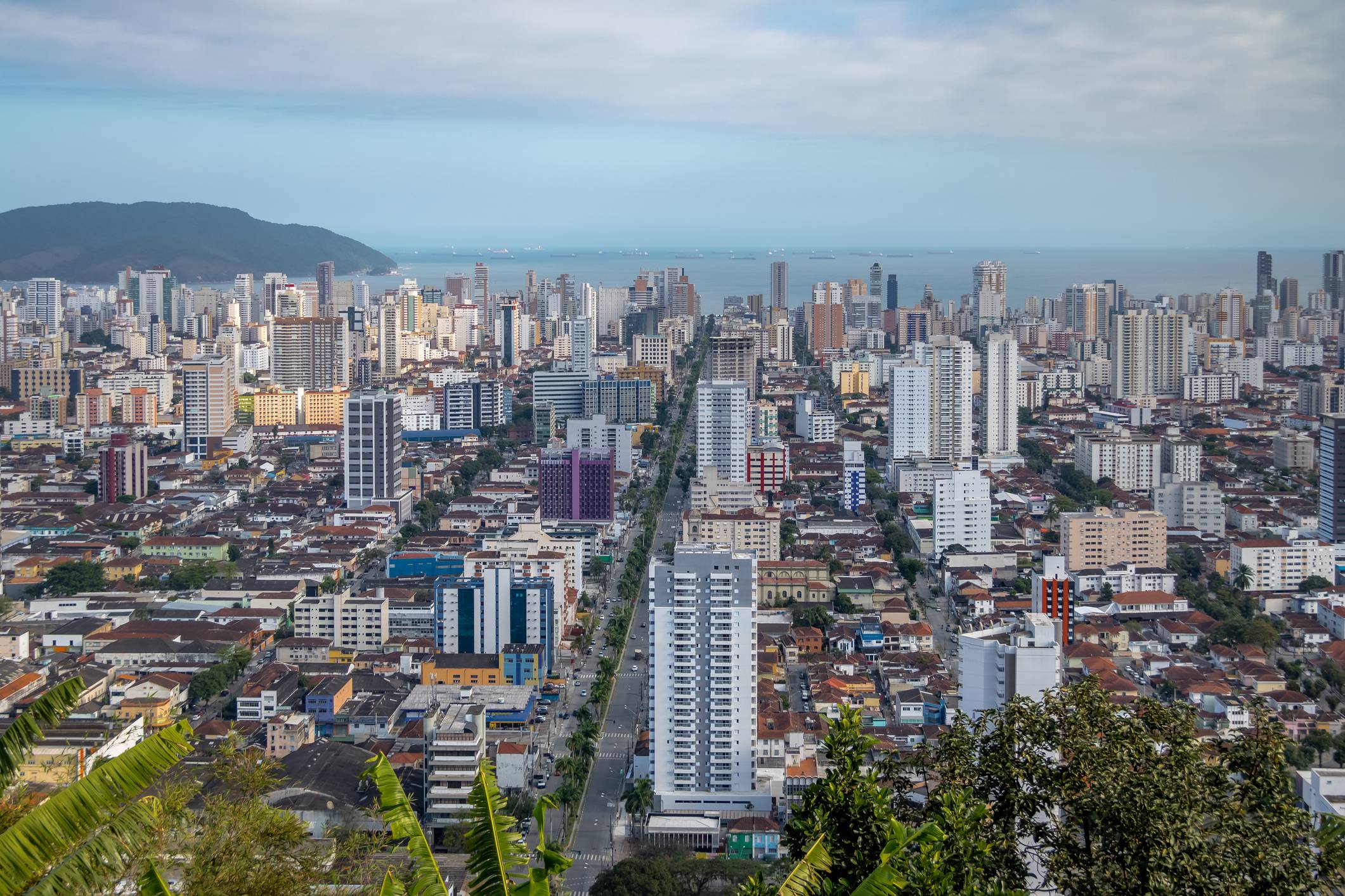 Vista aérea de Santos com prédios, avenida principal, mar com navios e montanha ao fundo.