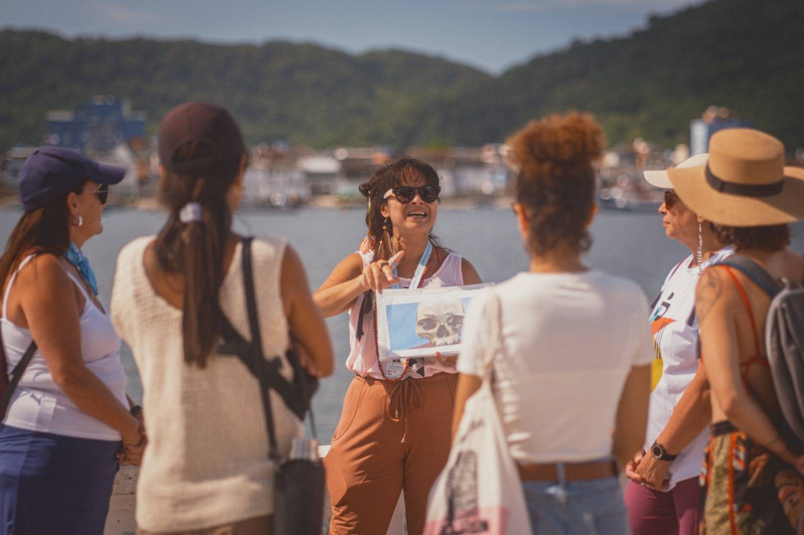 Guia turística sorrindo de óculos de sol mostra imagem de crânio a grupo de mulheres em passeio pela orla com montanhas ao fundo.