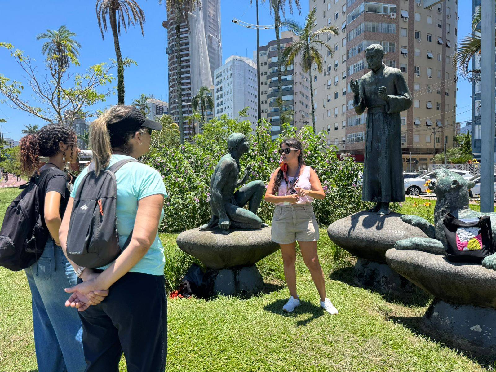 Mulher guia turística explicando sobre estátuas em um jardim urbano com edifícios e palmeiras ao fundo.