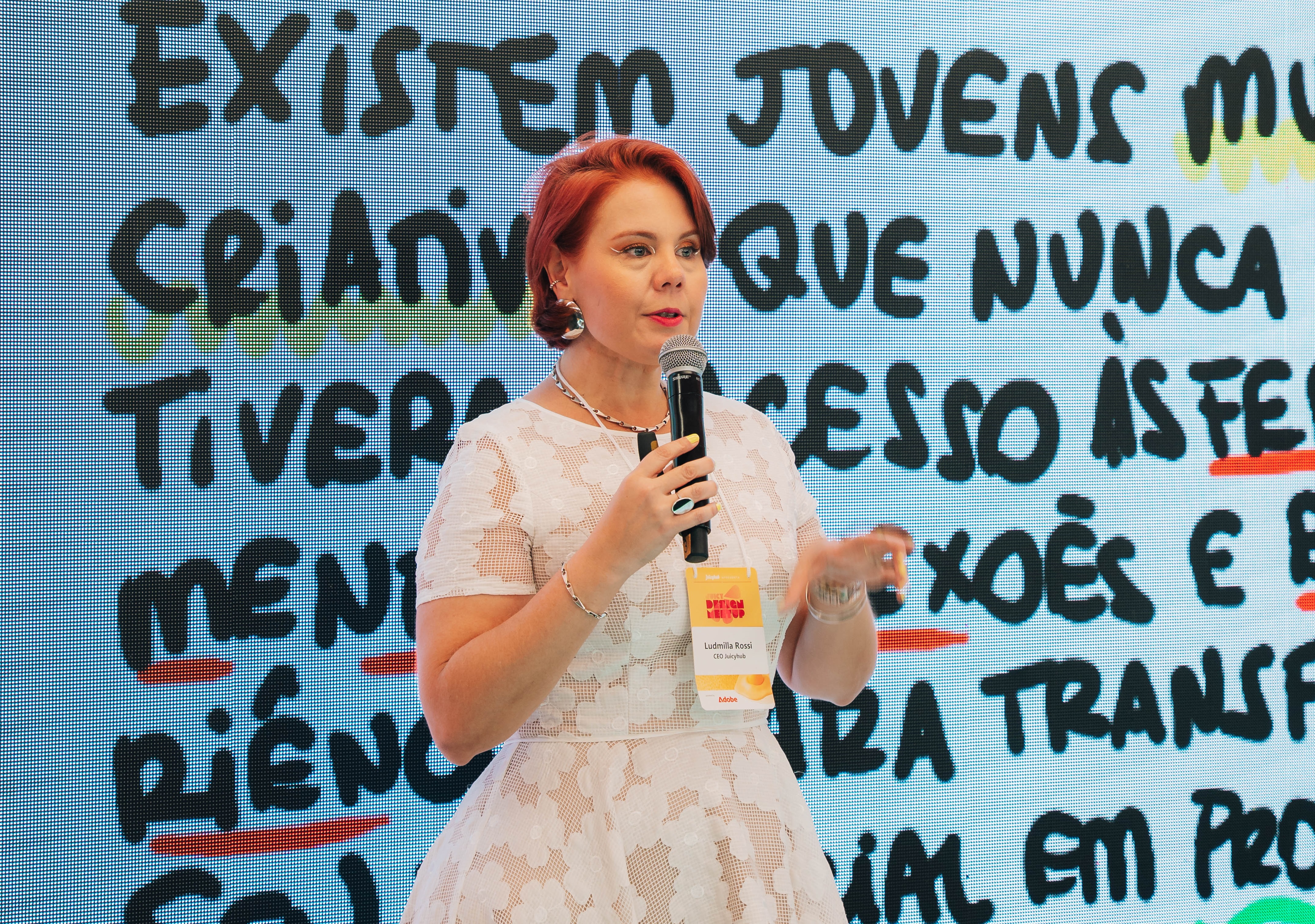 Ludmilla Rossi da Juicyhub apresentando em evento, segurando um microfone, com um telão LED de fundo exibindo texto sobre jovens e criatividade.