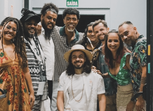 Grupo diverso de dez músicos e artistas sorrindo para a câmera, alguns com dreadlocks e chapéu, ao lado de uma placa de 'SAÍDA'.