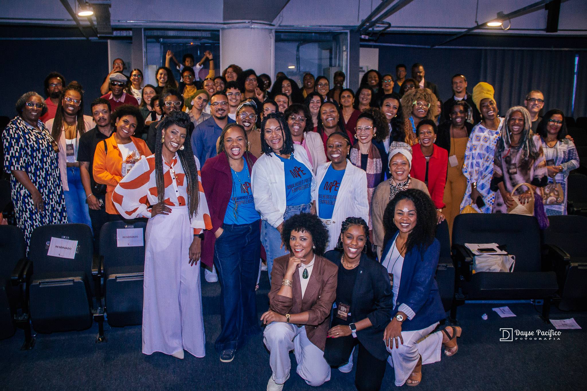 Grande grupo de mulheres e homens negros sorrindo em um evento, com algumas usando camisetas azuis e outras ajoelhadas à frente.
