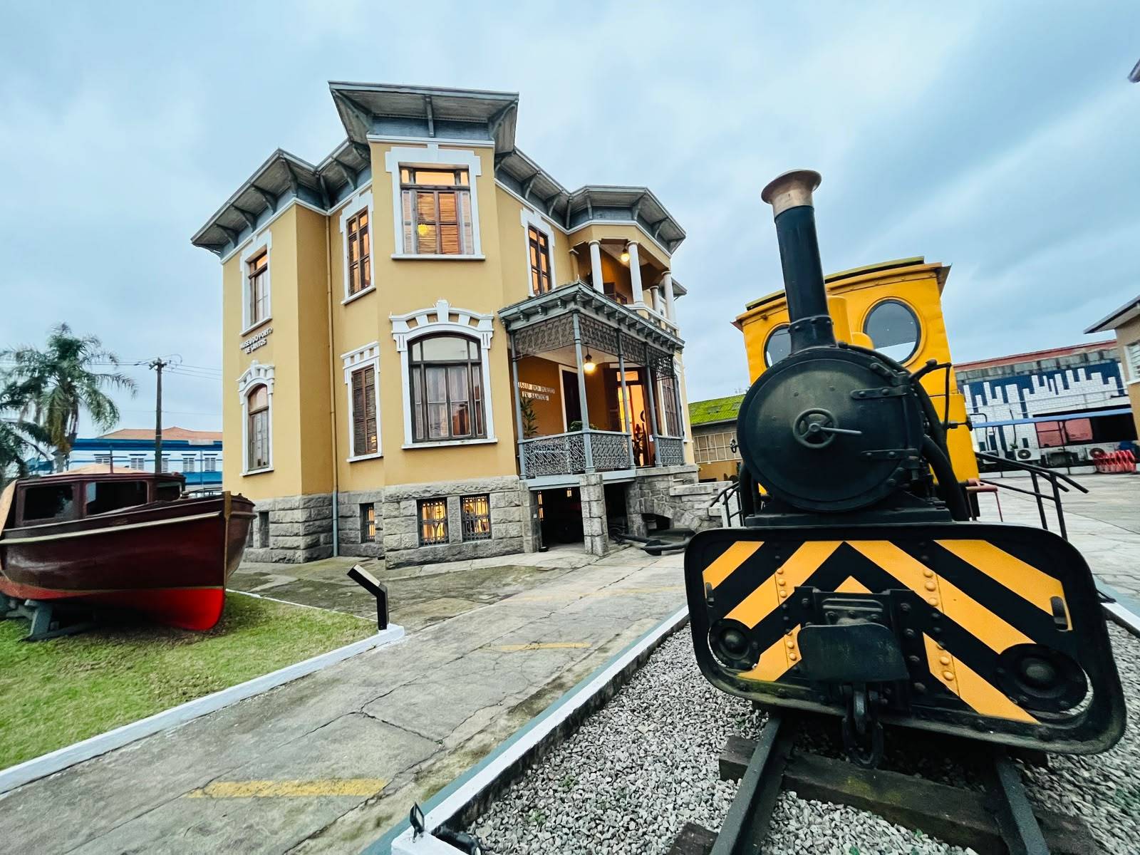 Museu do Porto de Santos, um edifício histórico amarelo com barco de madeira e locomotiva antiga em primeiro plano sob céu nublado.