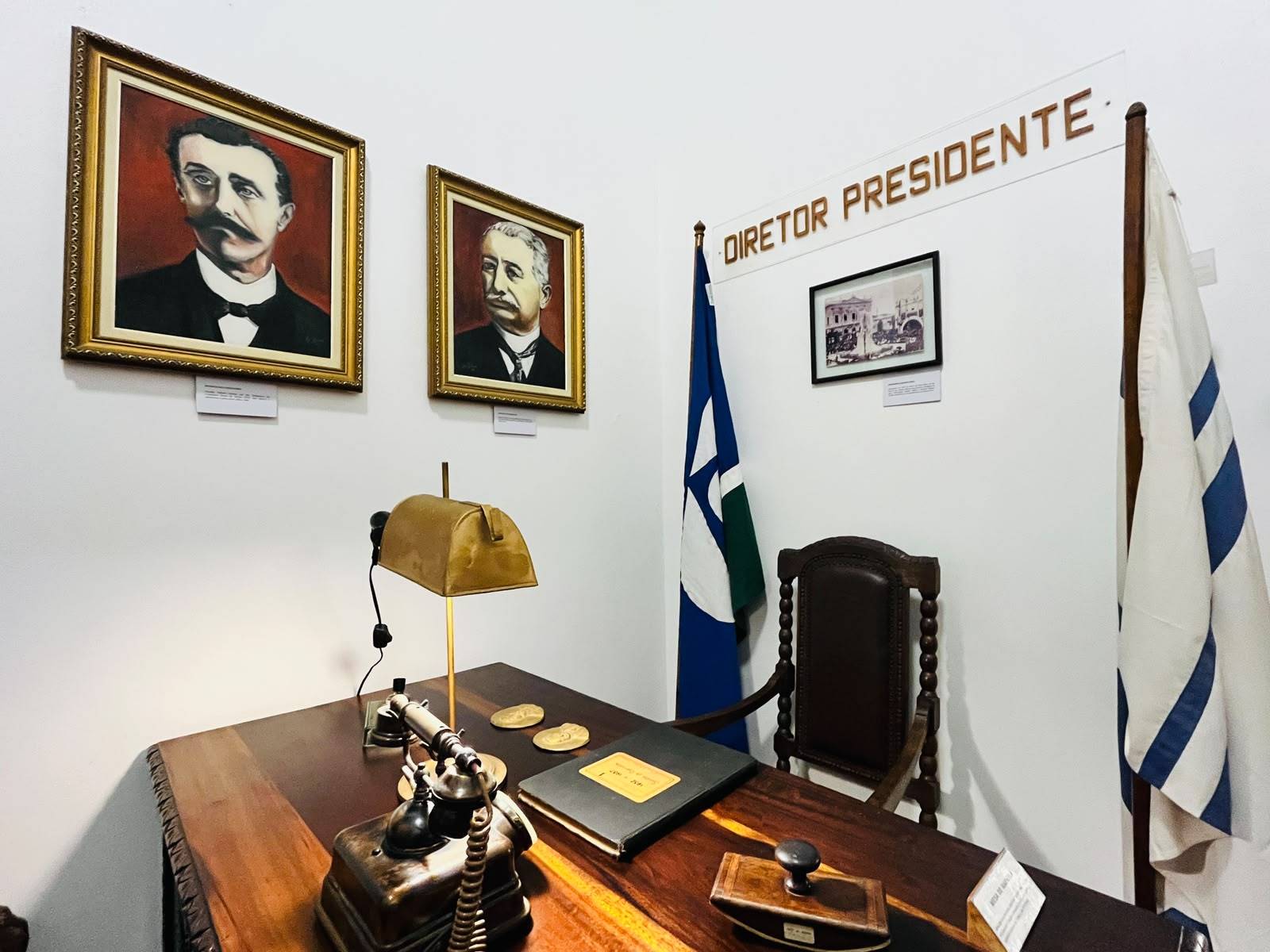 Exposição de escritório histórico com mesa de madeira, telefone vintage, luminária e retratos de ex-diretores. Duas bandeiras flanqueiam uma cadeira.