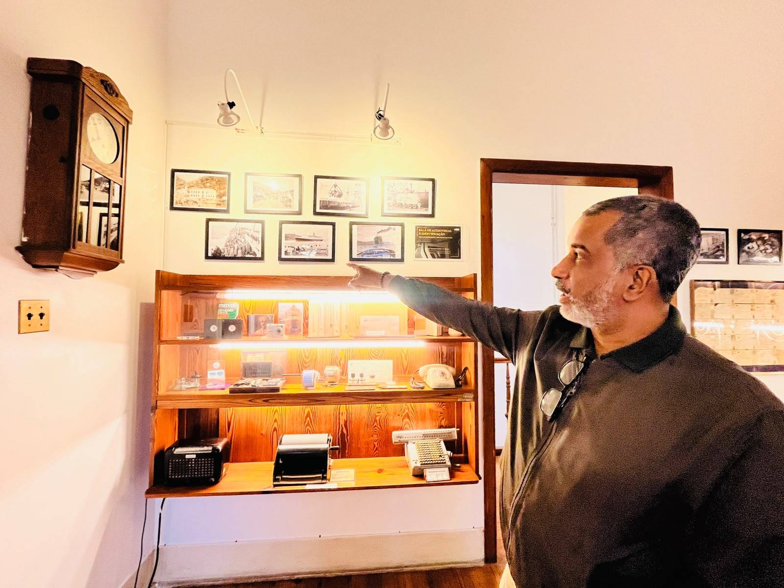 Homem de perfil com barba e óculos apontando para uma estante de madeira iluminada com objetos antigos e fotos em preto e branco.