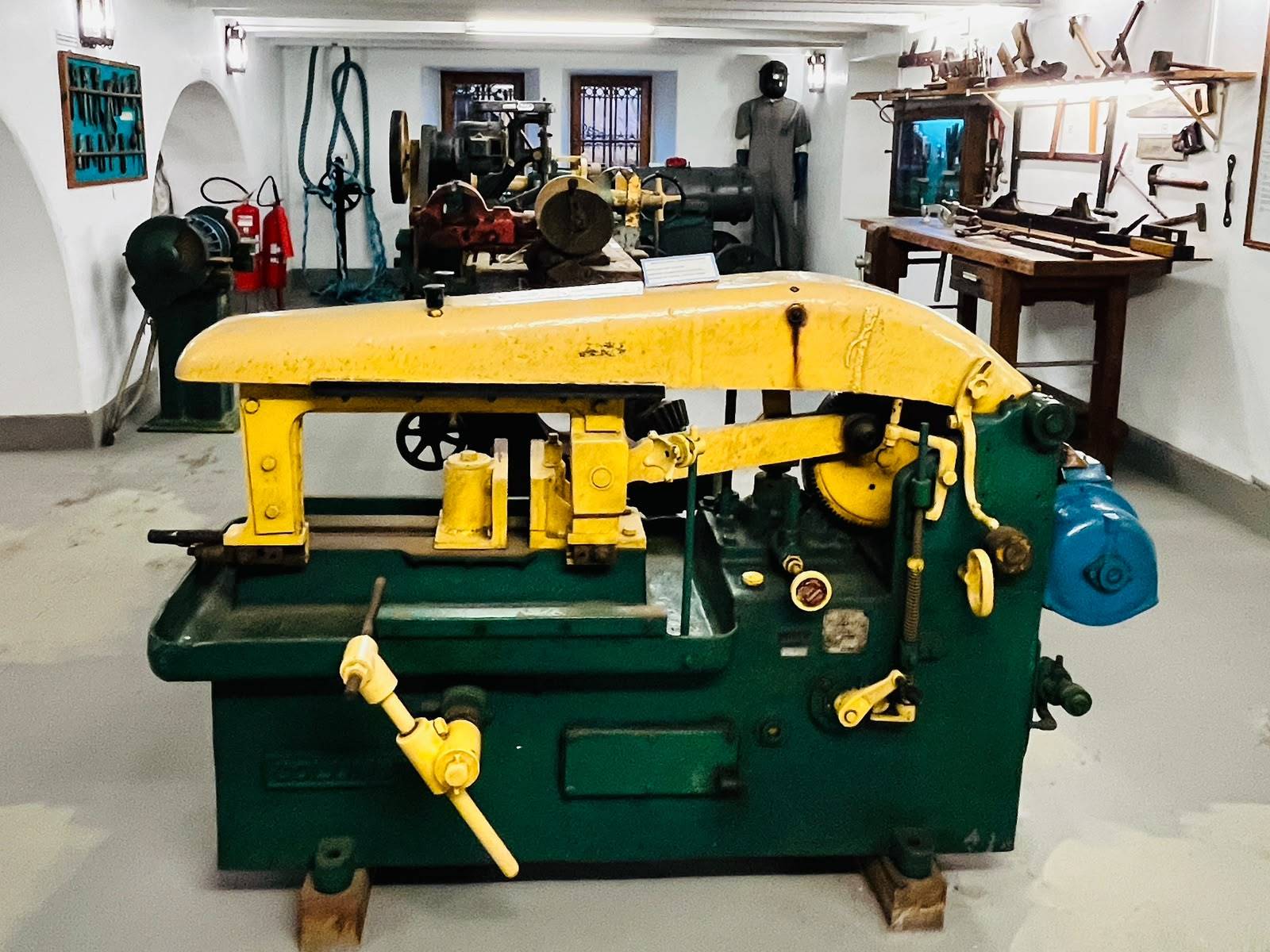 Antiga máquina industrial amarela e verde em primeiro plano, com diversas ferramentas e maquinário de oficina ao fundo.