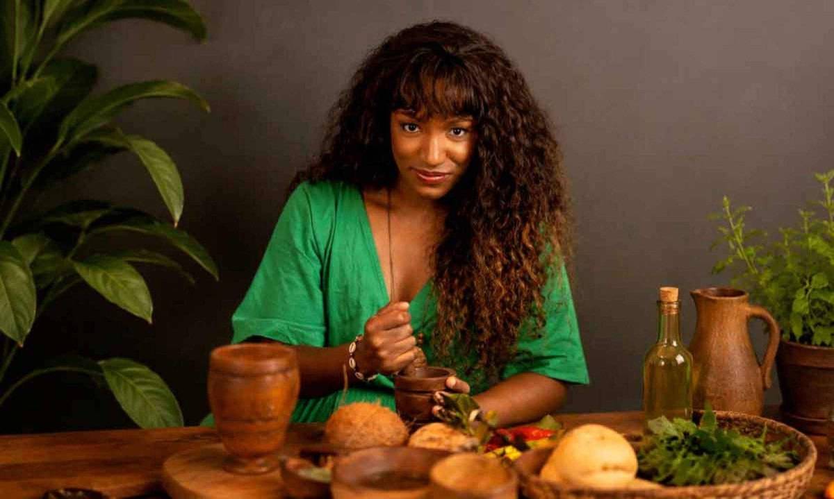Mulher de cabelos cacheados esmagando ervas em pilão, com ingredientes frescos e plantas na mesa.