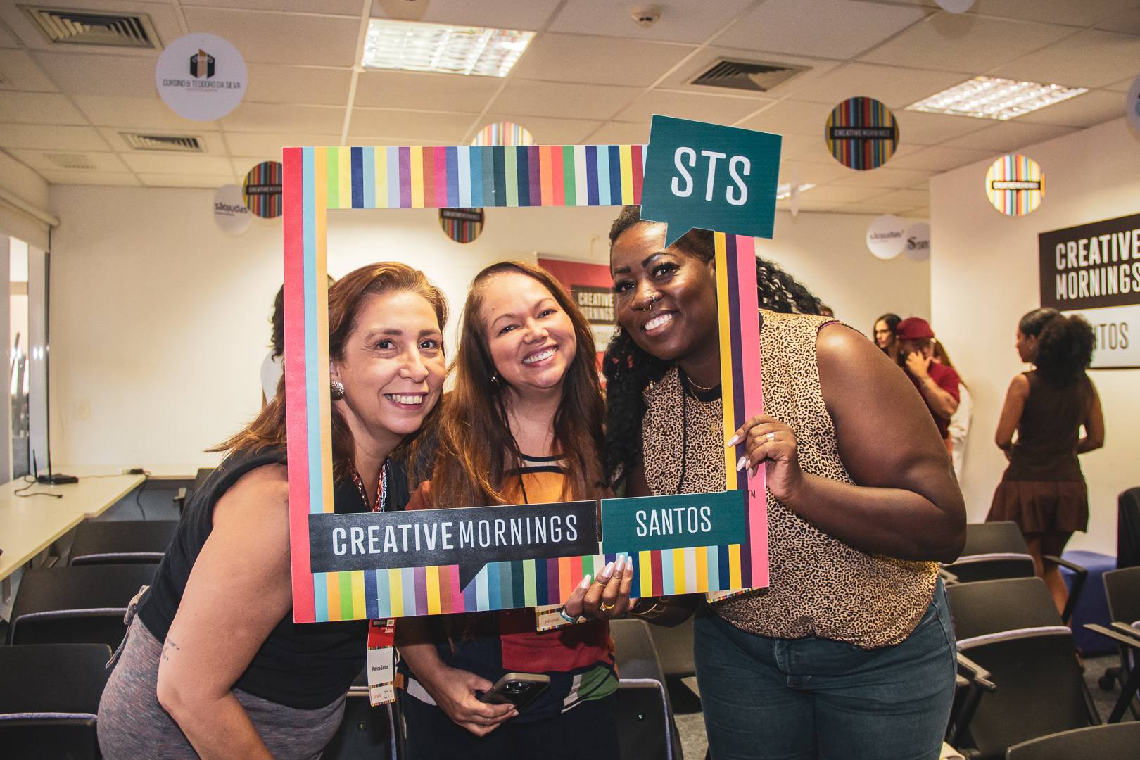 Três mulheres sorrindo em um evento Creative Mornings Santos, posando com moldura colorida personalizada do evento.