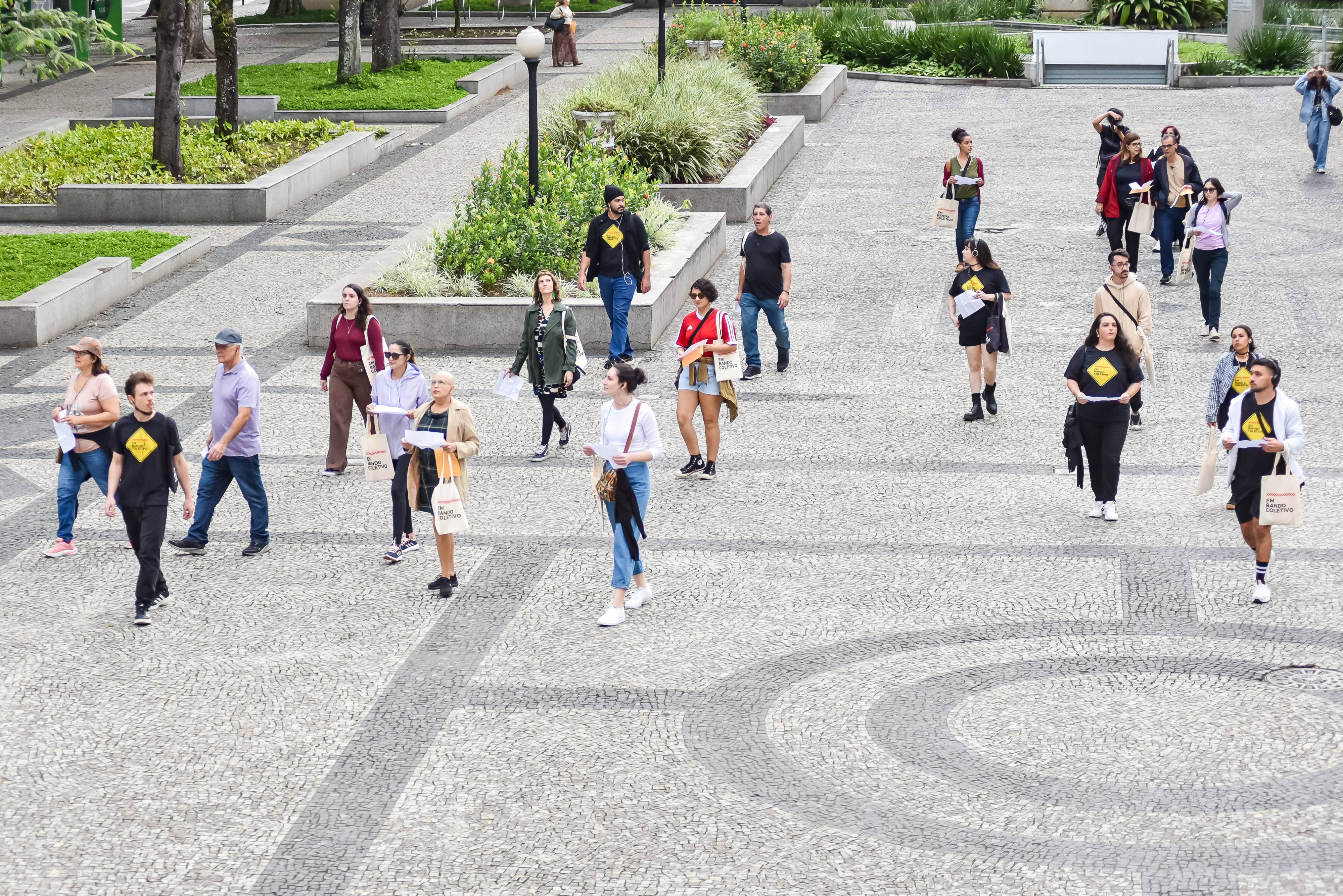 Pessoas caminhando em praça de paralelepípedos, muitas com camisetas pretas e bolsas brancas 'EM BANDO COLETIVO'.