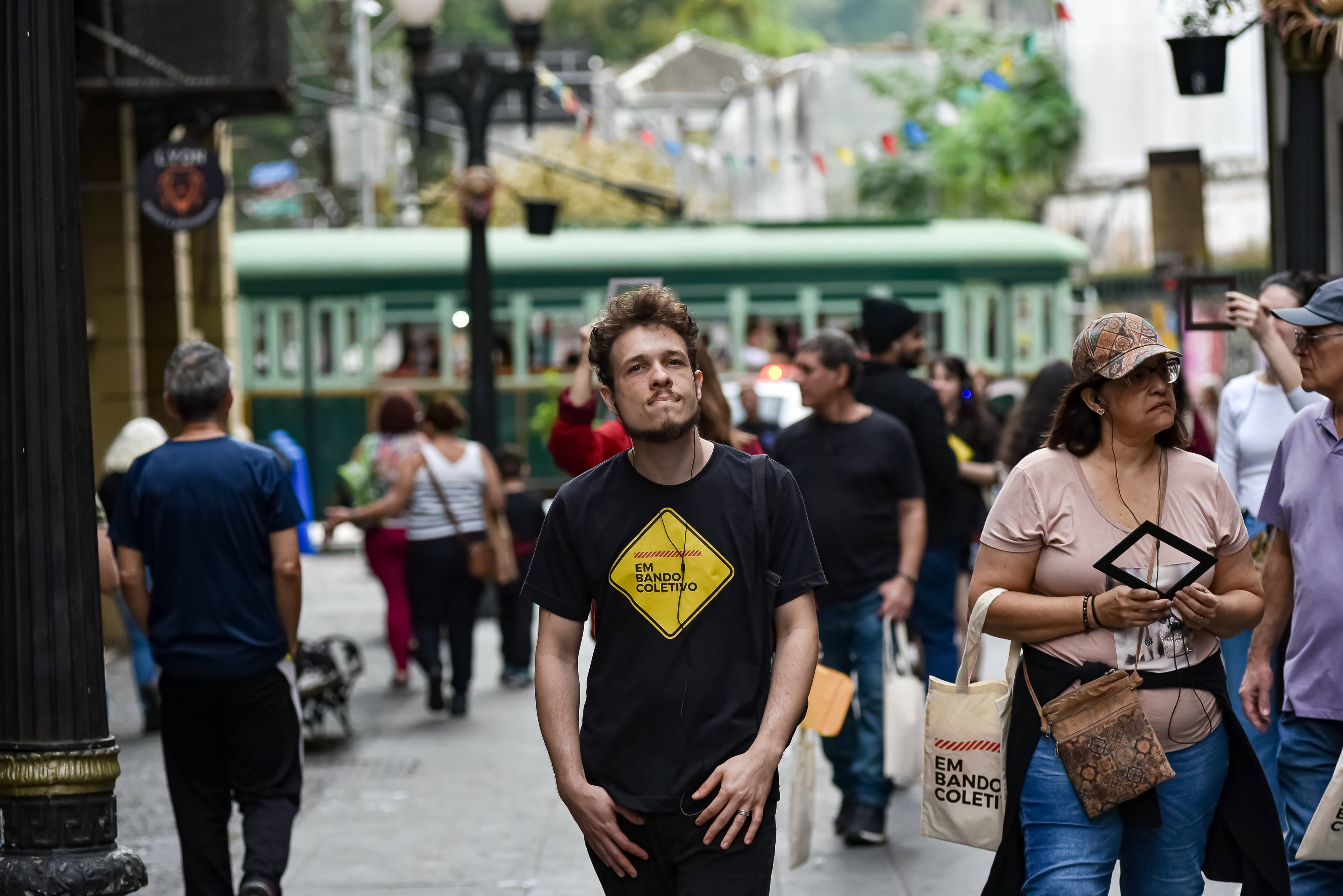Jovem e mulher usando camisetas 'EM BANDO COLETIVO' em rua movimentada com bonde verde ao fundo.