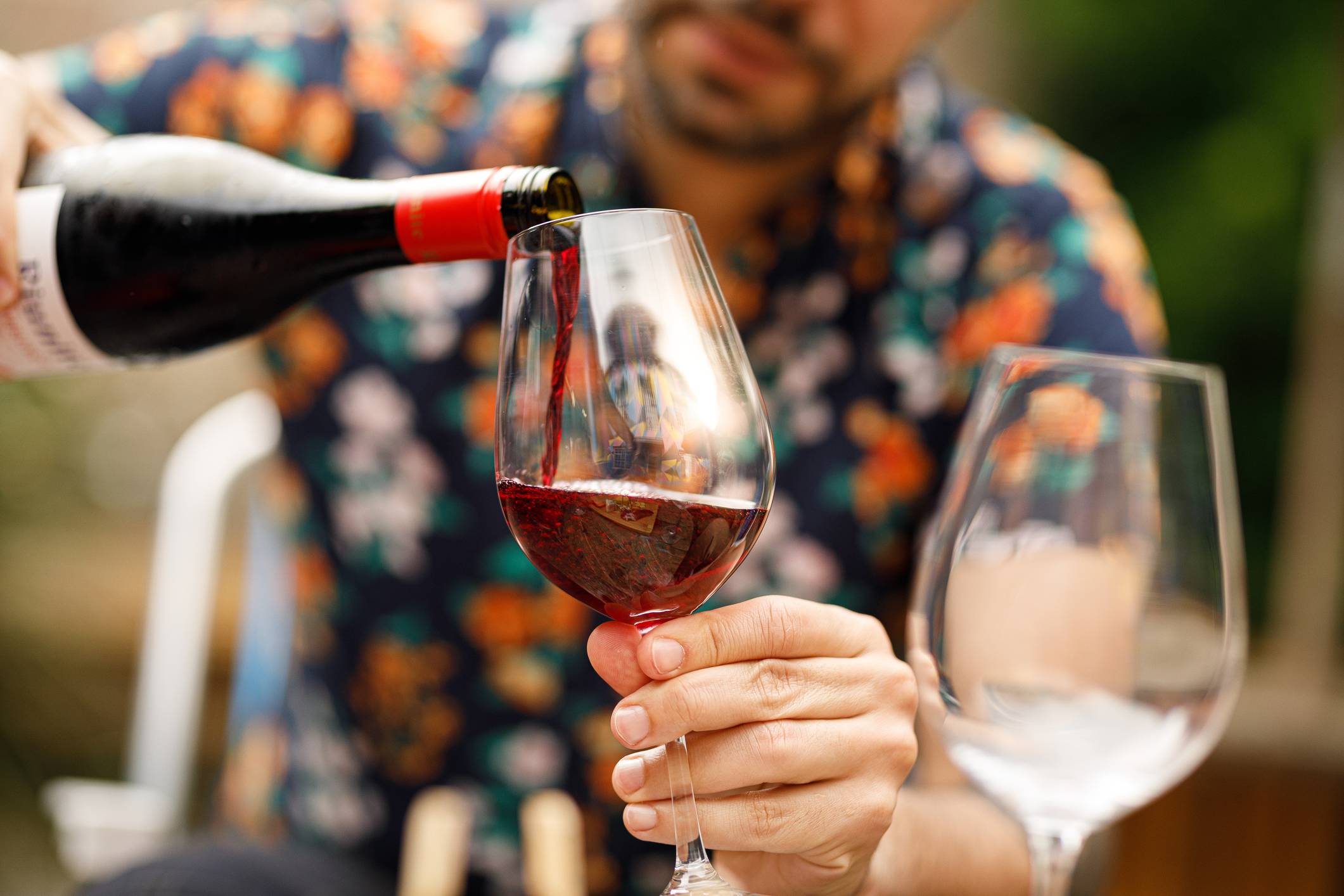 Homem servindo vinho tinto de uma garrafa em uma taça, com outra taça vazia ao lado, em foco.