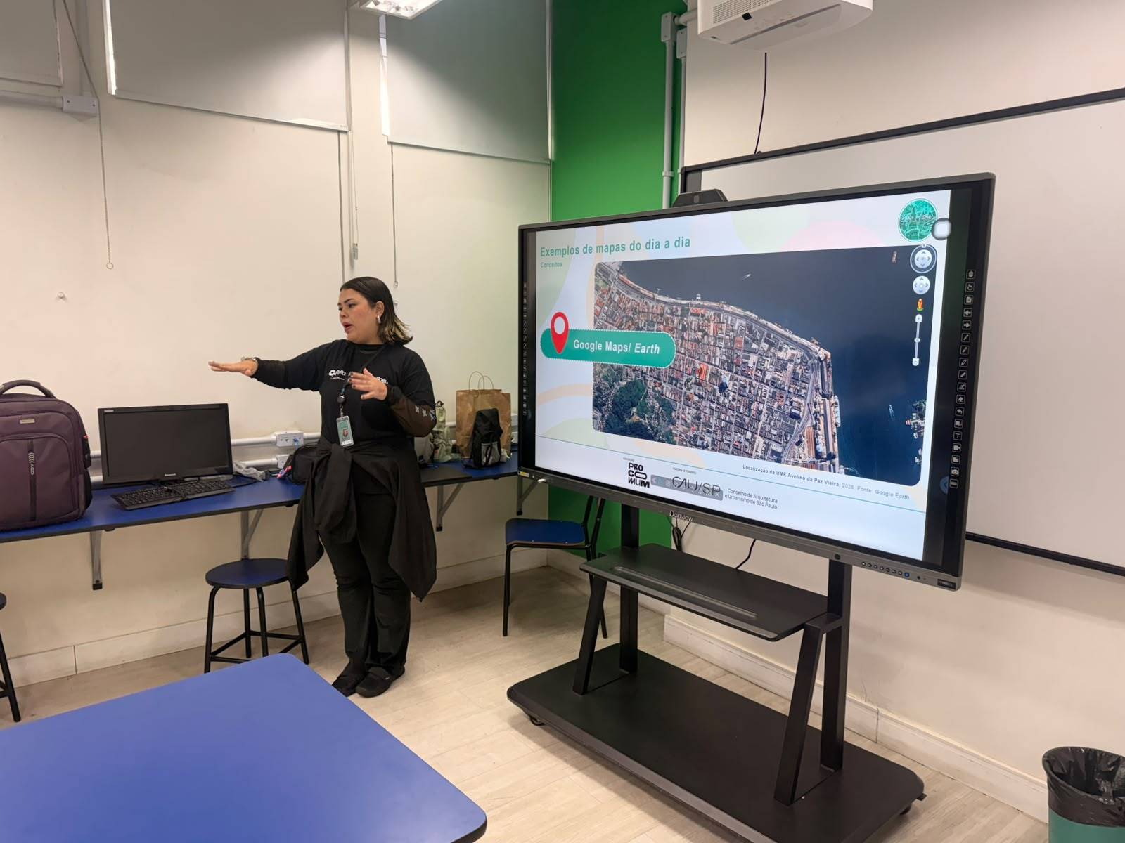 Mulher apresenta mapa aéreo do Google Maps em tela interativa para uma audiência em sala de aula.