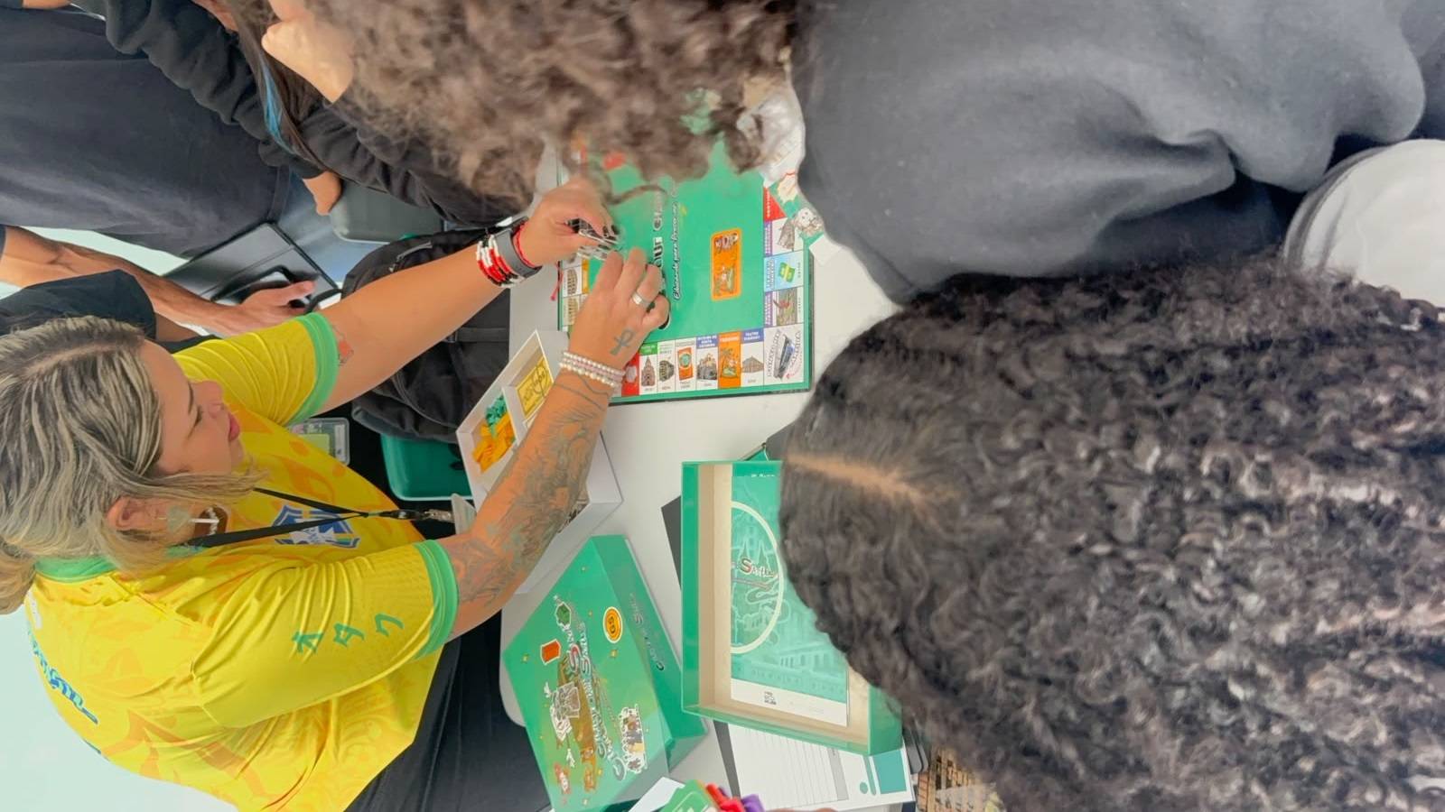 Mulher em camisa amarela joga board game verde com temática de cidade, outras pessoas observam.
