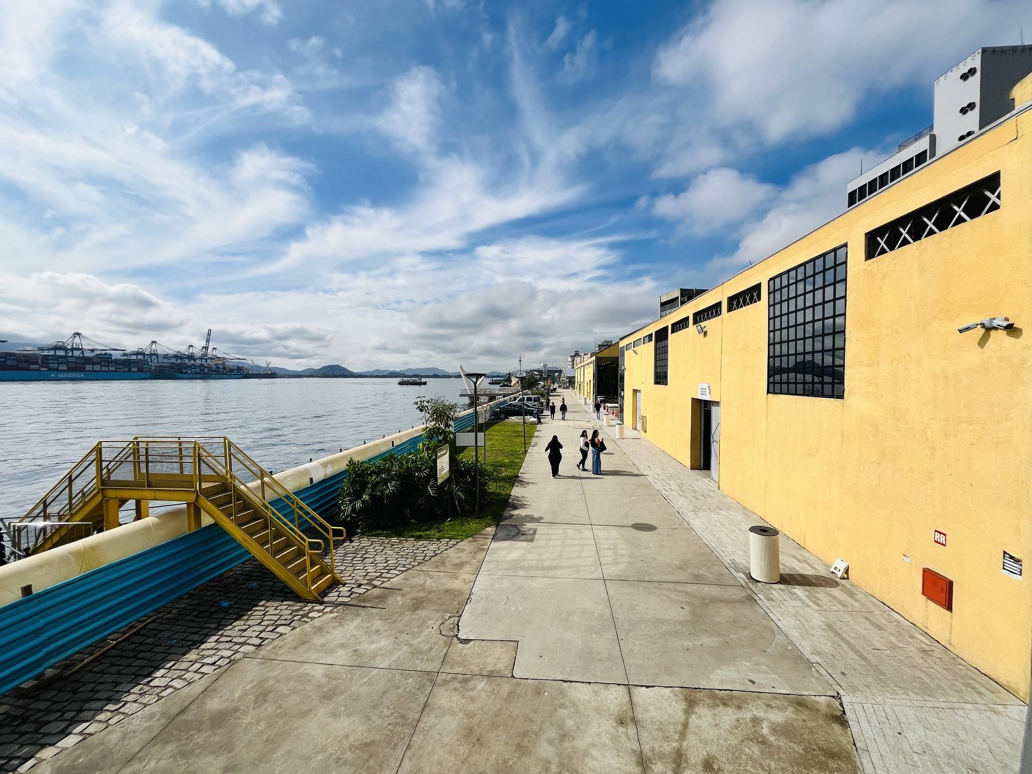 Passeio público com edifício amarelo, cais, navios cargueiros e montanhas sob céu azul com nuvens