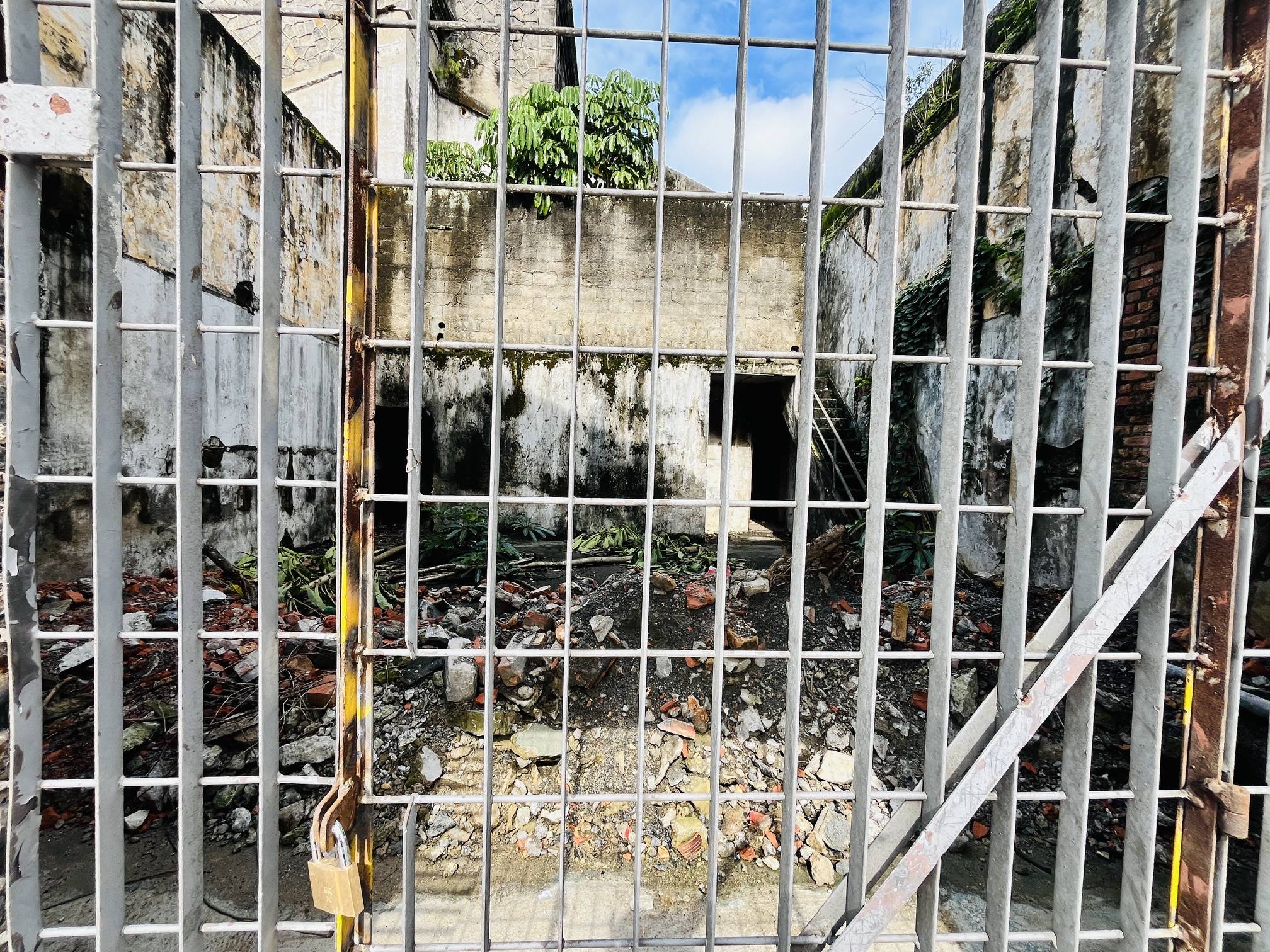 Edifício abandonado e dilapidado visto através de um portão de metal trancado, com entulho no chão e vegetação crescendo.