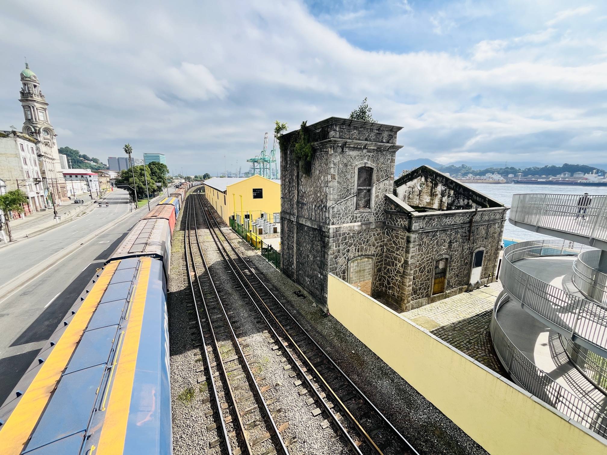Trilhos de trem com vagões azuis e amarelos, edifícios de pedra e porto com guindastes e navios ao fundo, em Santos.
