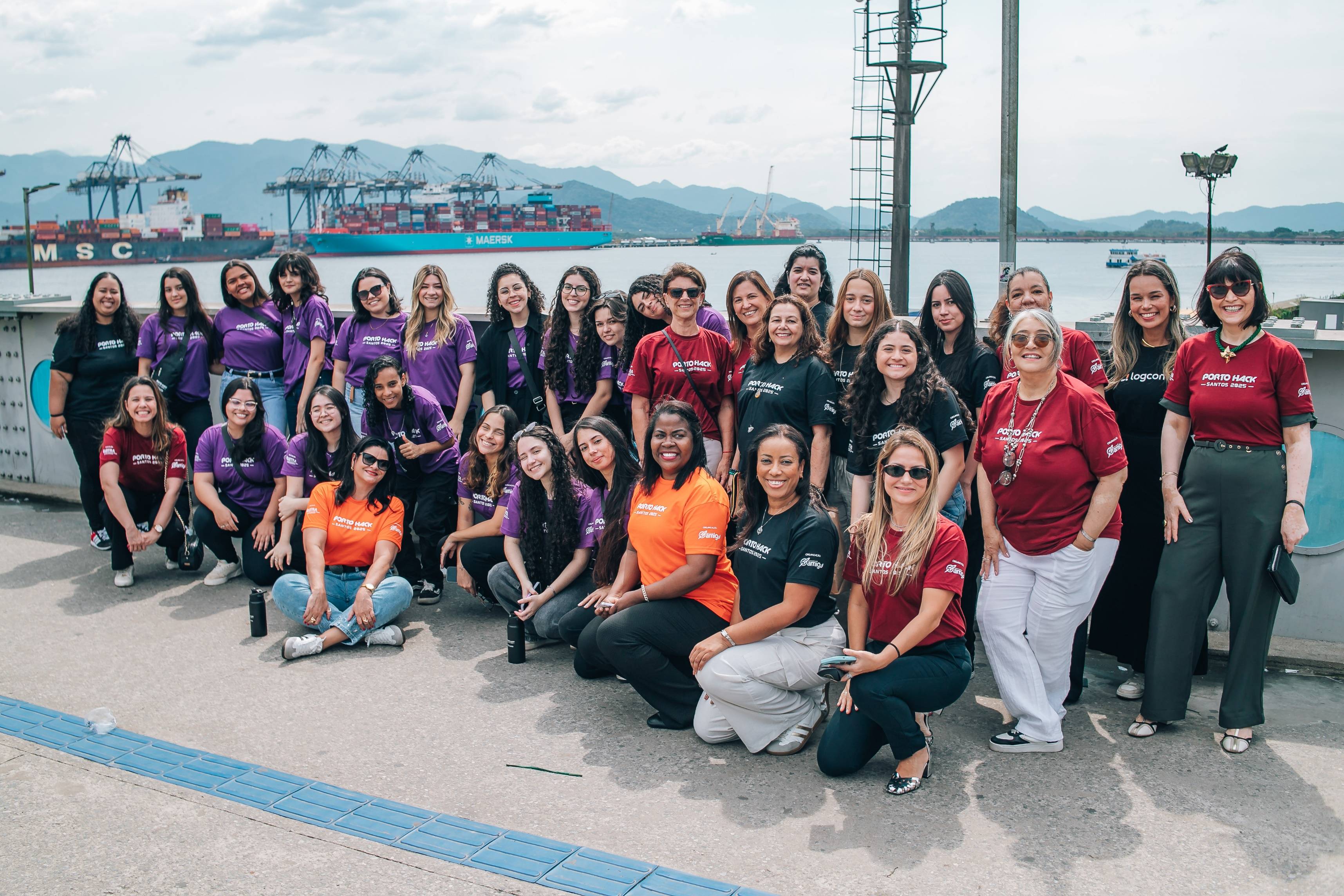 Diverse group of women in 'Porto Hack Santos 2025' shirts posing at a port with container ships and mountains.