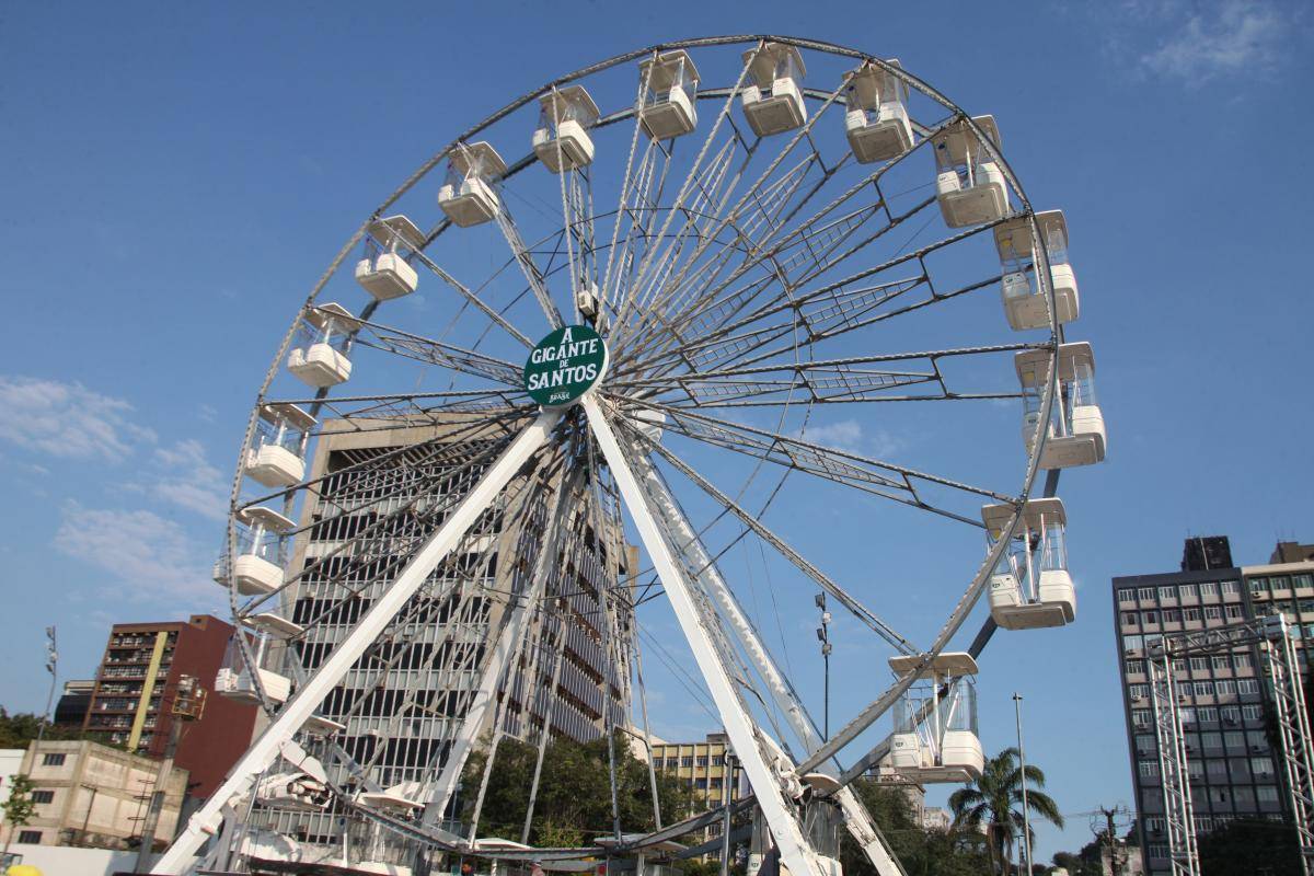 Roda-gigante branca com várias cabines contra um céu azul claro, exibindo um letreiro verde central 'A Gigante de Santos'