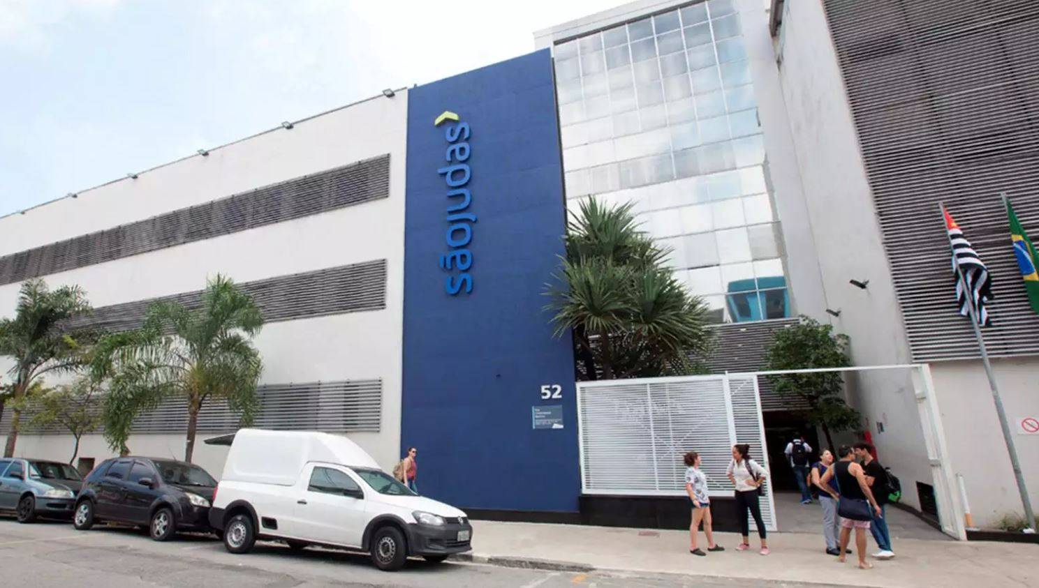 Fachada principal da Universidade São Judas com letreiro azul, carros estacionados e pessoas na calçada, sob céu claro.