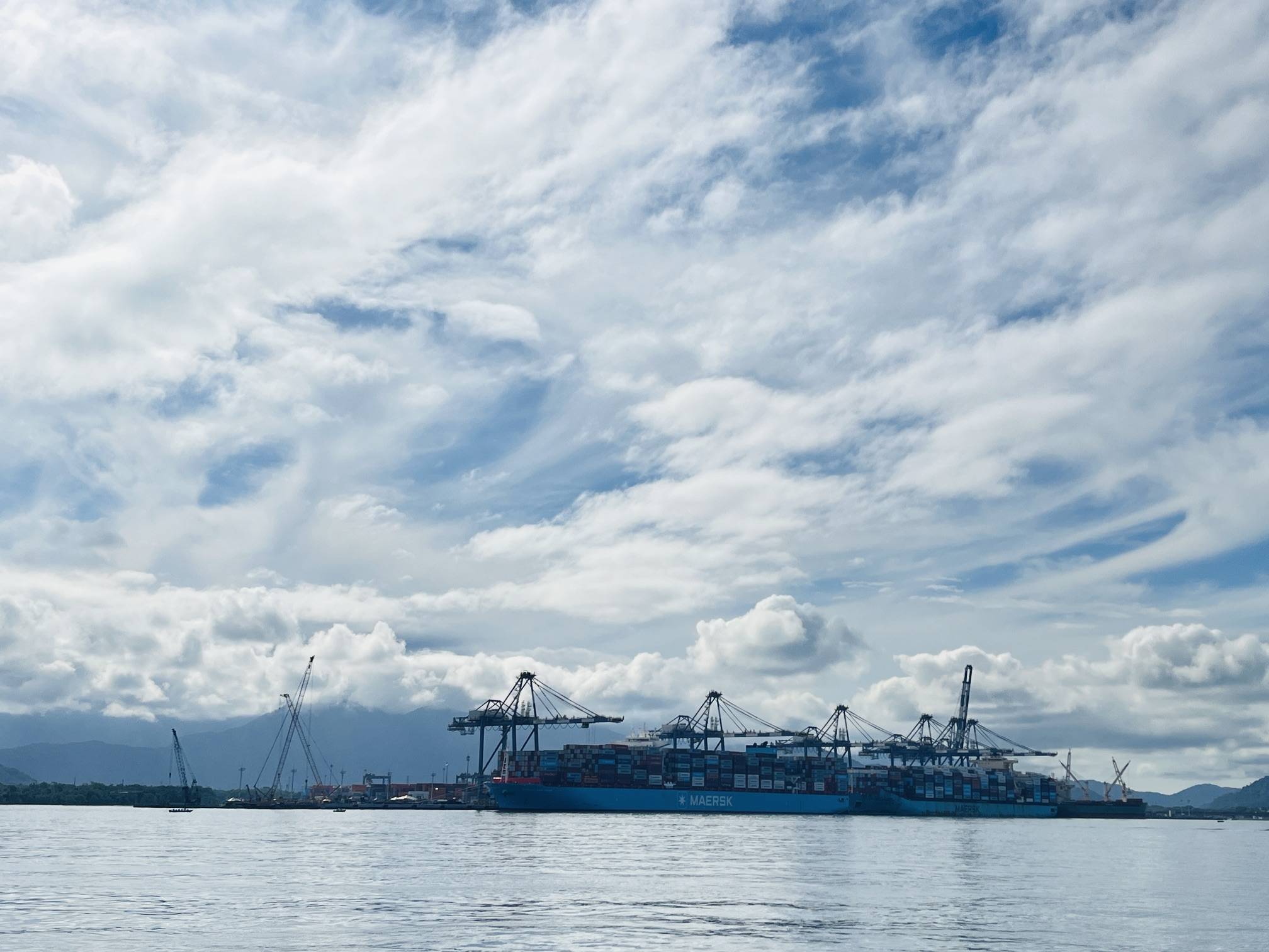 Navios cargueiros da Maersk em porto industrial com guindastes de contêineres, montanhas e céu nublado sobre a água calma.