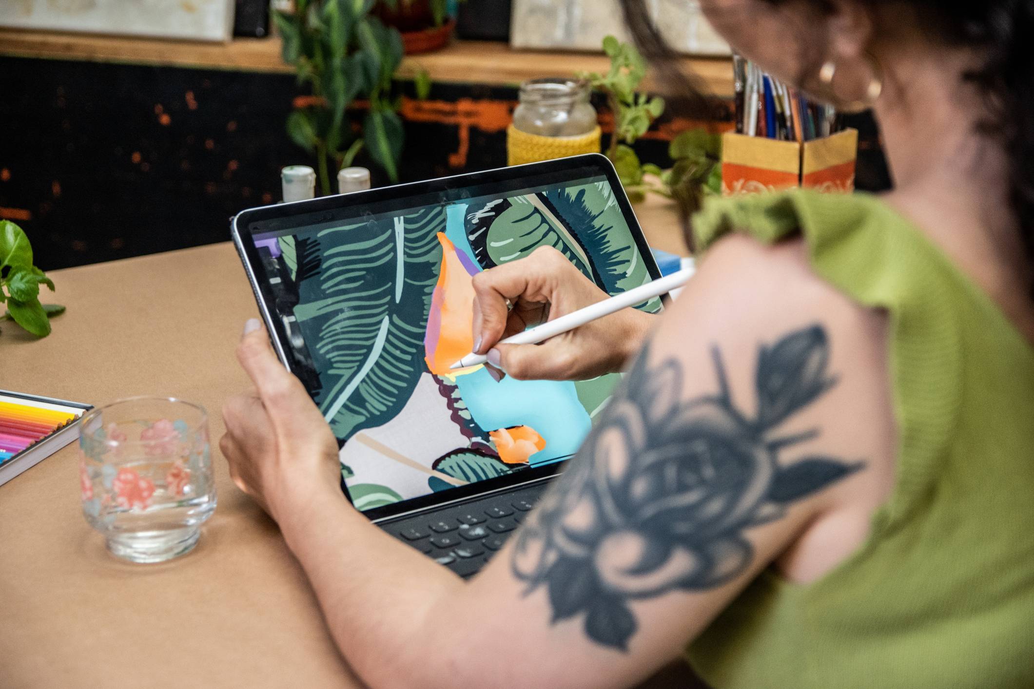 Pessoa desenhando digitalmente em tablet com caneta stylus, criando arte tropical e abstrata, com tatuagem de rosa no braço.