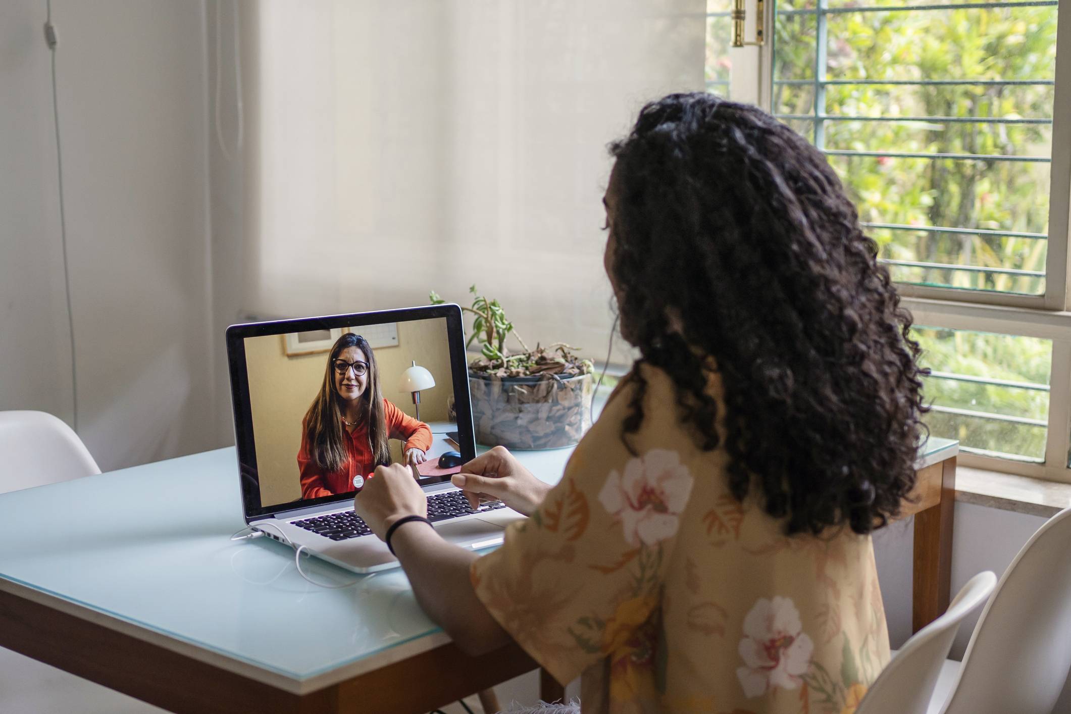 Pessoa com cabelo cacheado em videochamada no notebook, olhando para mulher de óculos na tela. Foco em trabalho remoto e conexão digital.