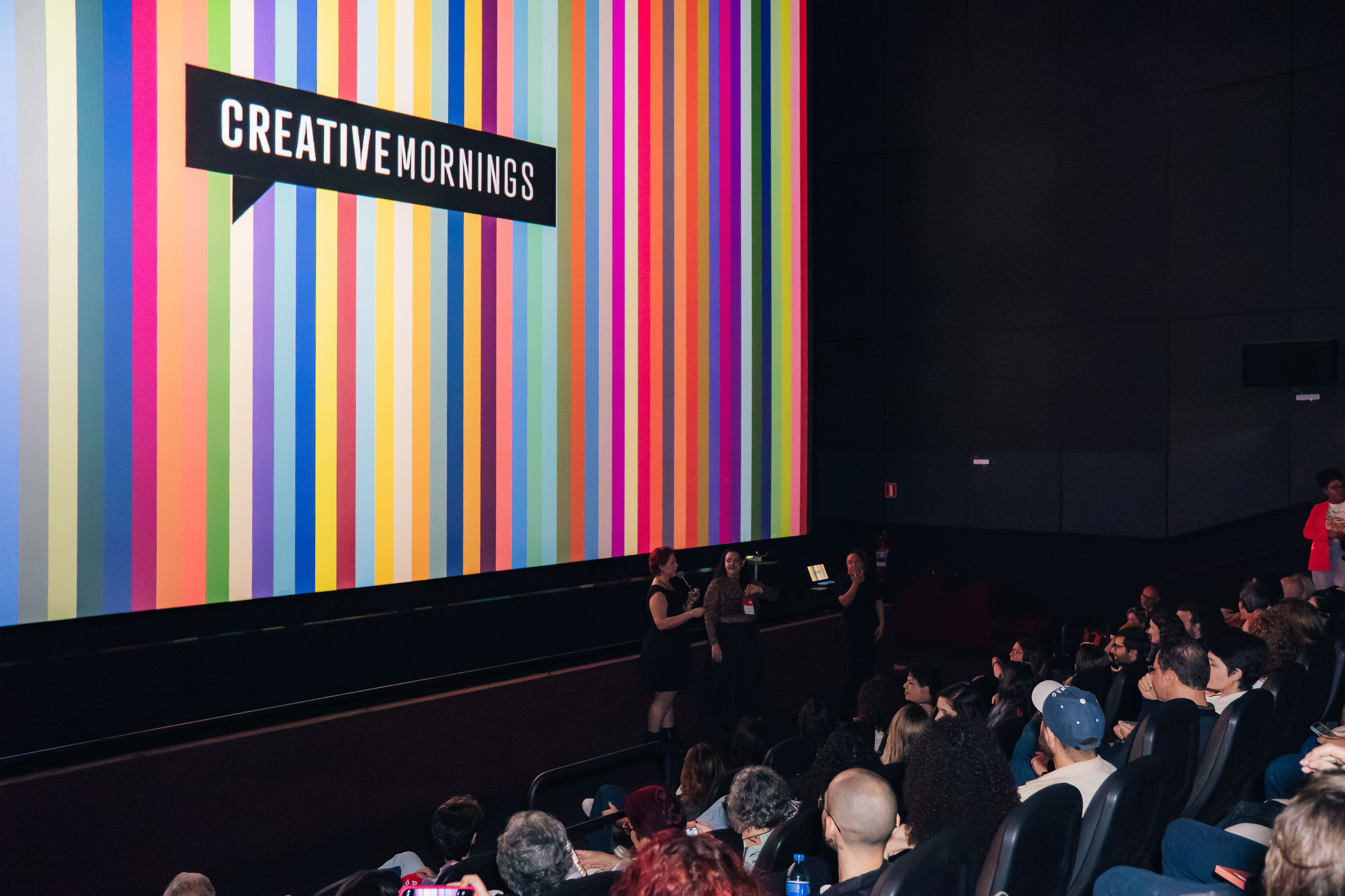 Palestrantes no palco à frente de uma tela grande com listras coloridas verticais e o logo 'CREATIVEMORNINGS', com público sentado.