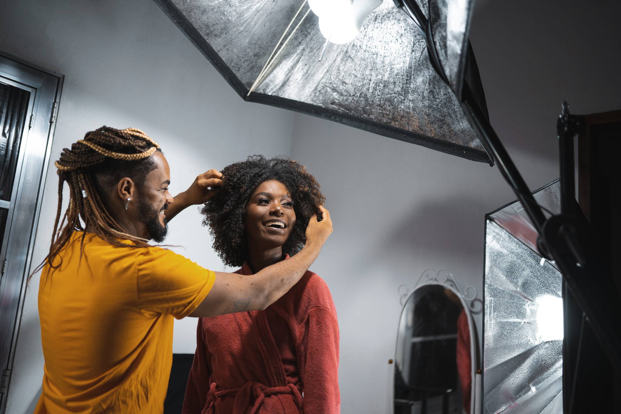 Um homem com tranças ajeita o cabelo cacheado de uma mulher sorridente em um estúdio com luzes softbox.