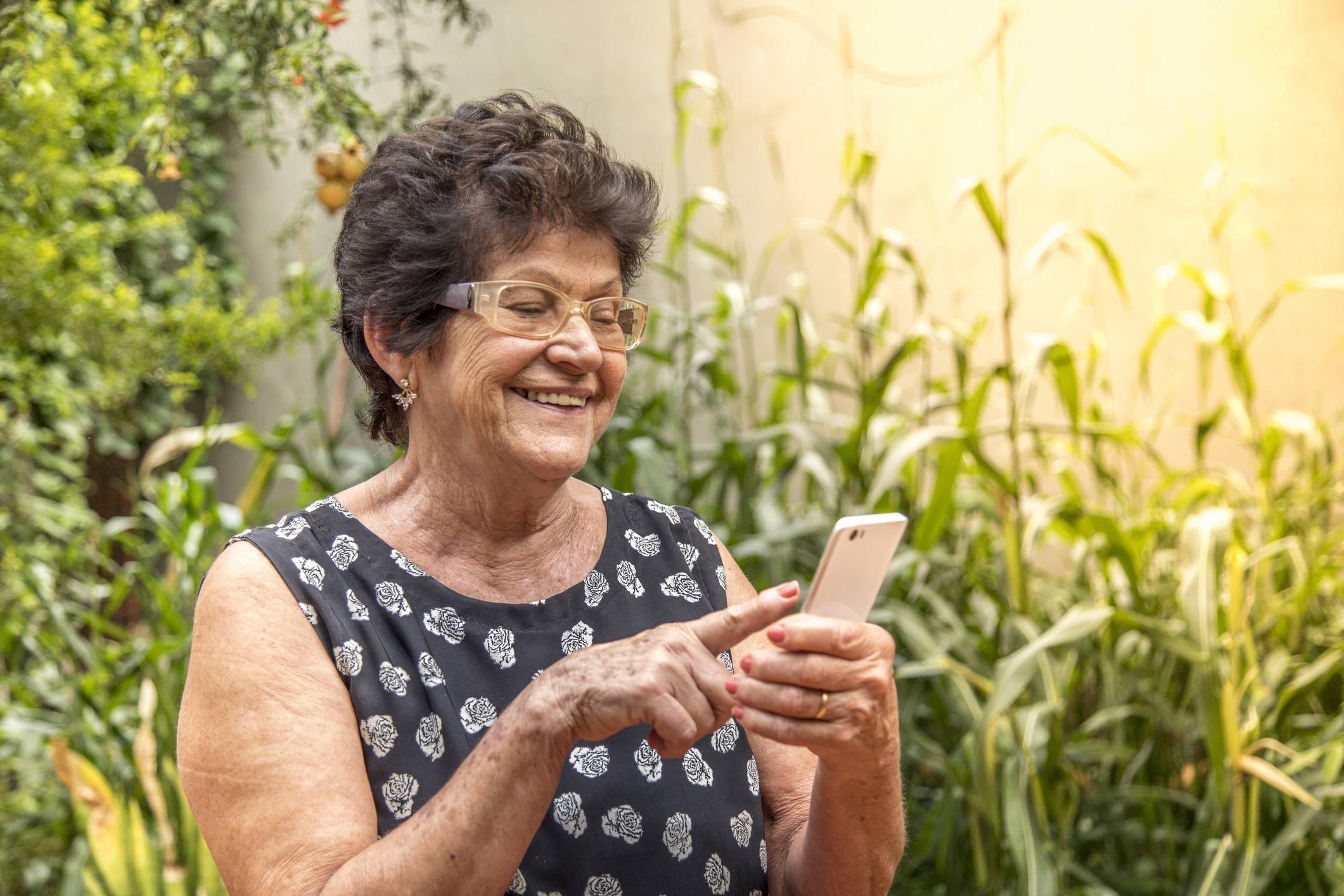 Mulher idosa sorridente de óculos, interagindo com smartphone branco em jardim ensolarado.